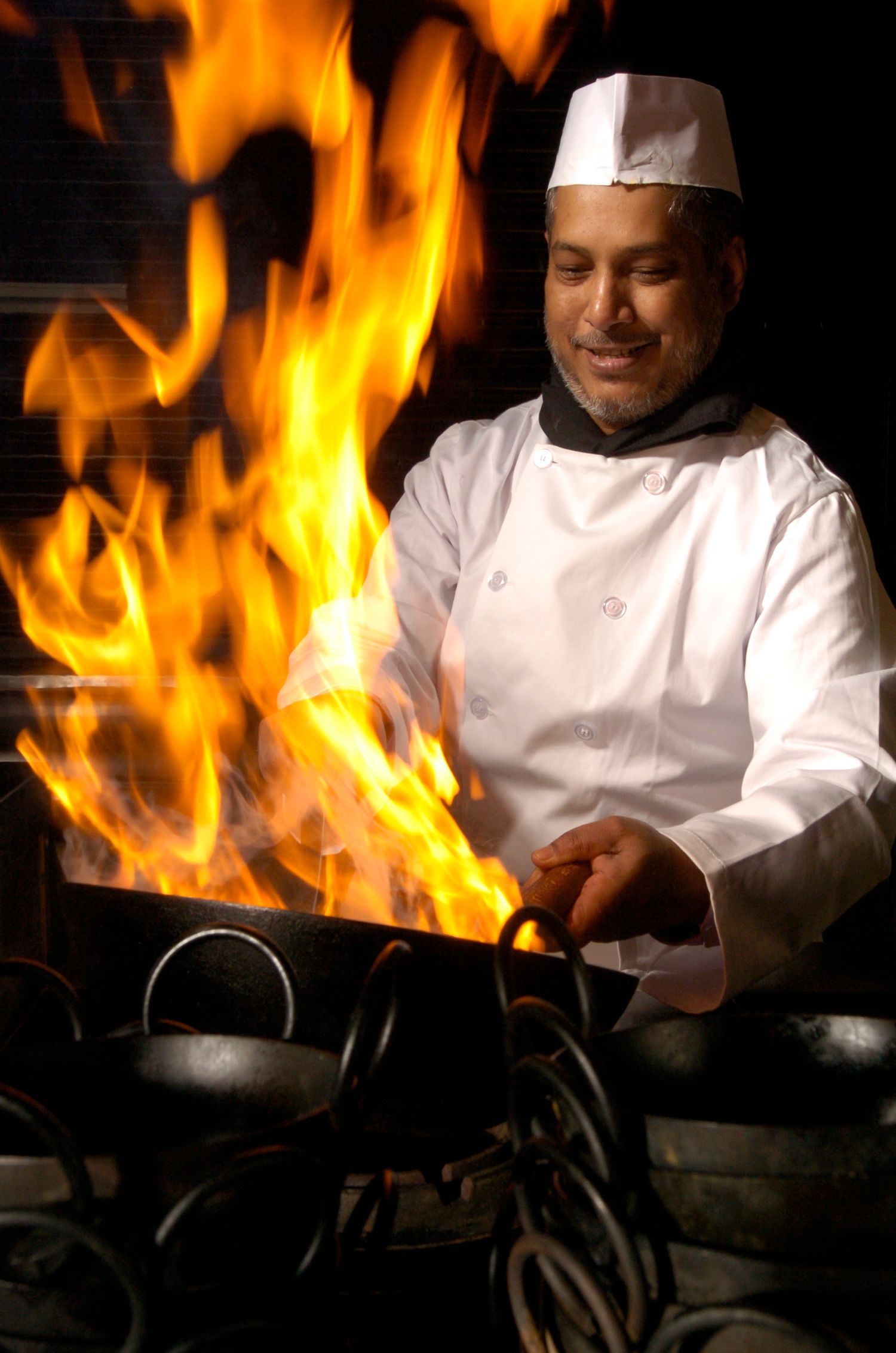 A smiling chef in white uniform and hat cooking over dramatic open flames in a professional kitchen.
