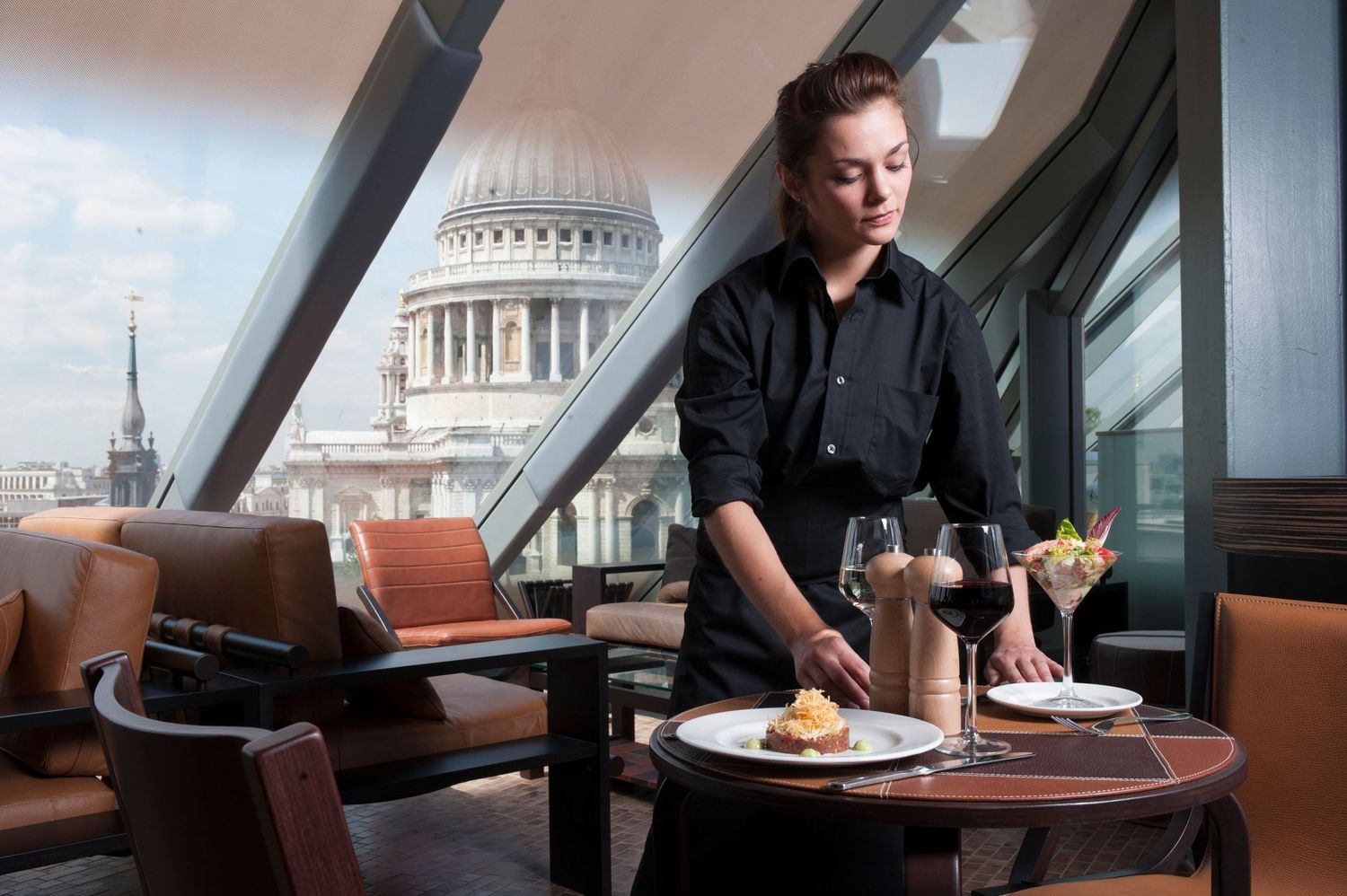 Waitress serving elegant dishes at a rooftop restaurant with St Paul's Cathedral dome visible through panoramic windows.