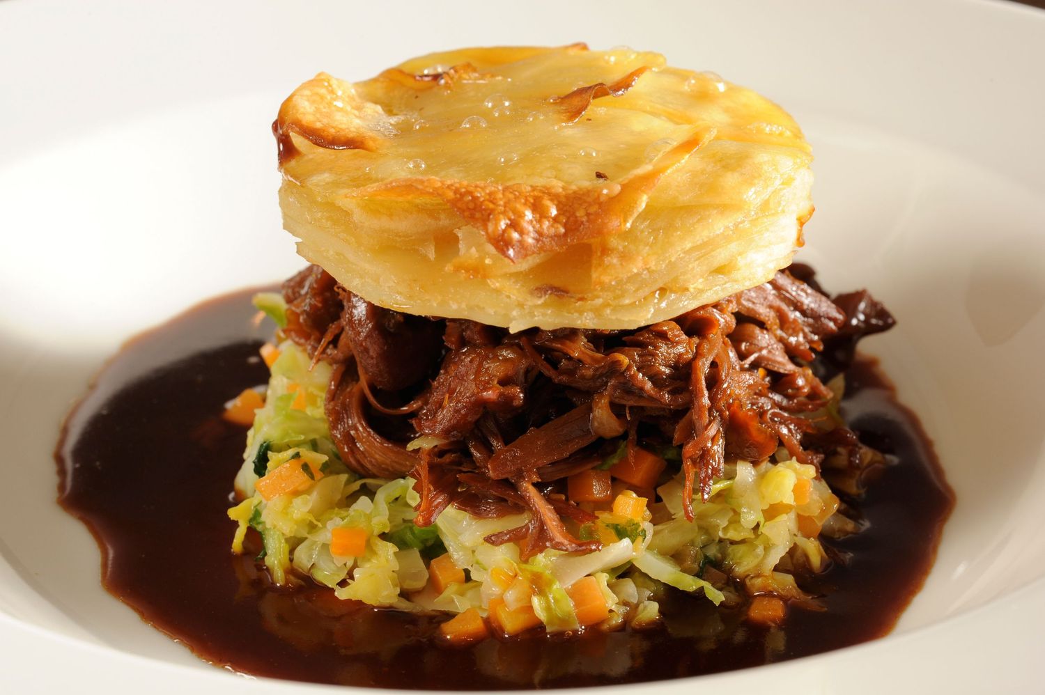 Slow-braised pulled beef served on vegetable hash with rich gravy, topped with a golden baked potato rosti on a white plate.