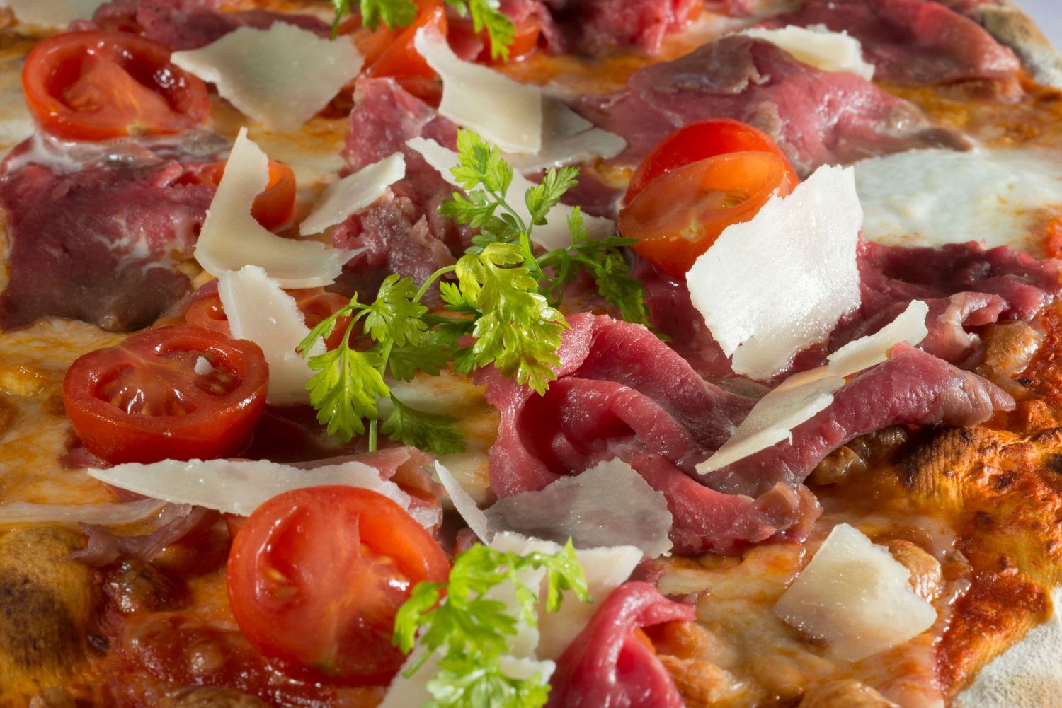 Close-up of a gourmet pizza topped with prosciutto, cherry tomatoes, shaved Parmesan, and fresh parsley.