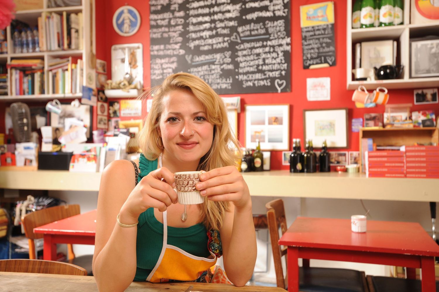 Young blonde woman smiling and holding a coffee cup in a vibrant red cafe with chalkboard menu.