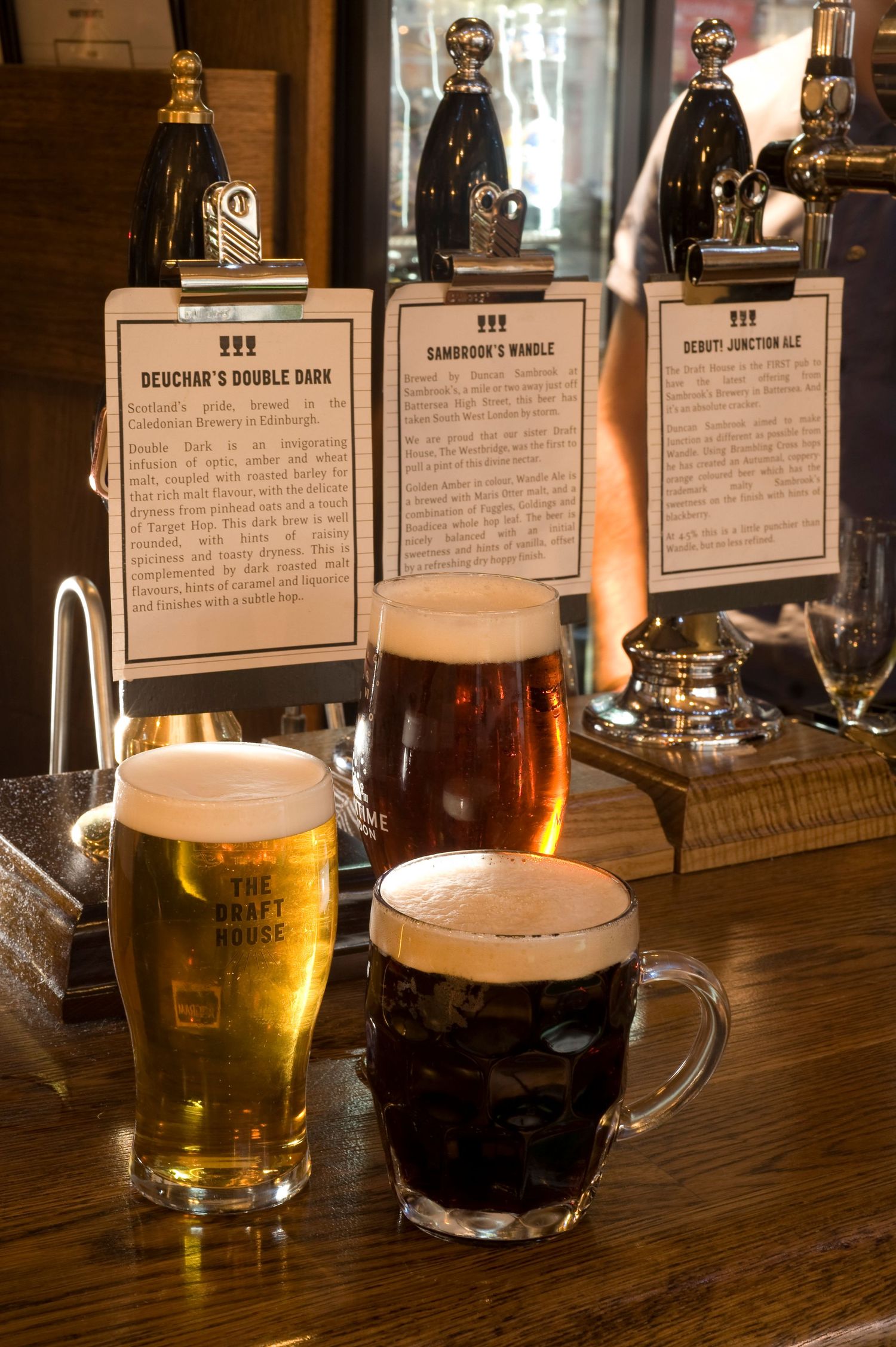Three craft beers on a pub bar, including a golden ale and dark stout, with tap handles and beer menu cards behind.