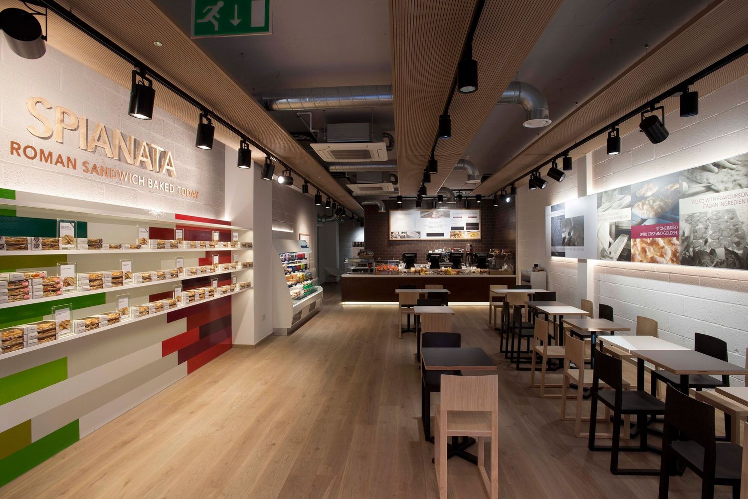 Interior of Spianata Roman Sandwich Bar with shelved products, wooden floors, track lighting, and modern dining area.