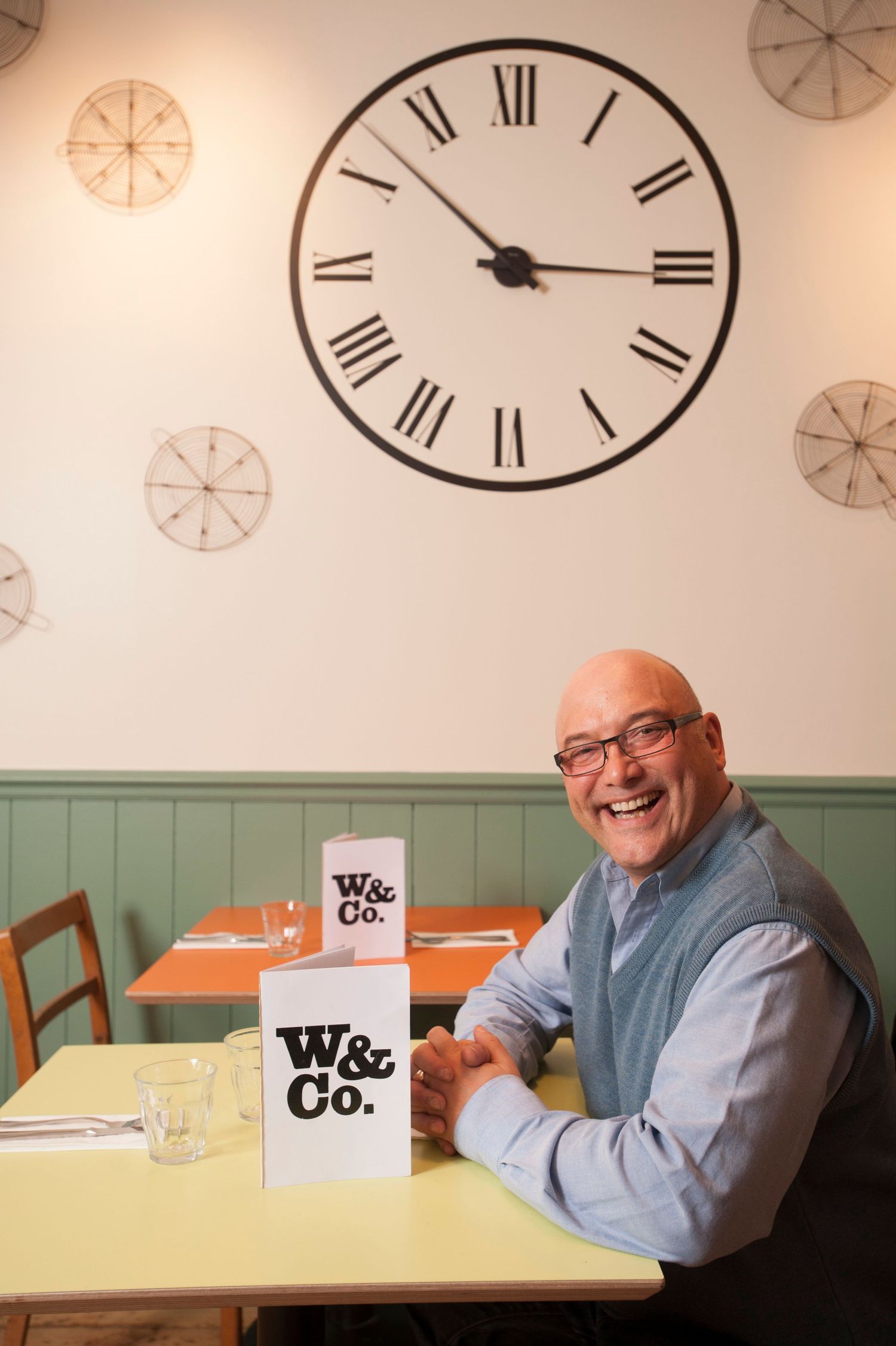 Smiling bald man sitting at a table in W&Co. restaurant with a large Roman numeral clock on the wall behind him.