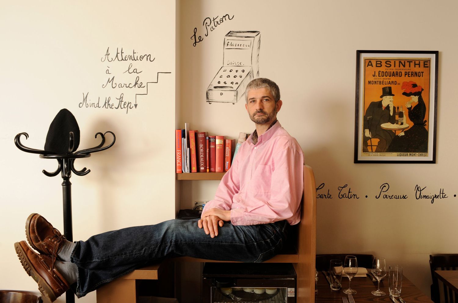 Man in pink shirt sits relaxed in a cafe with French wall art and an absinthe poster in the background.