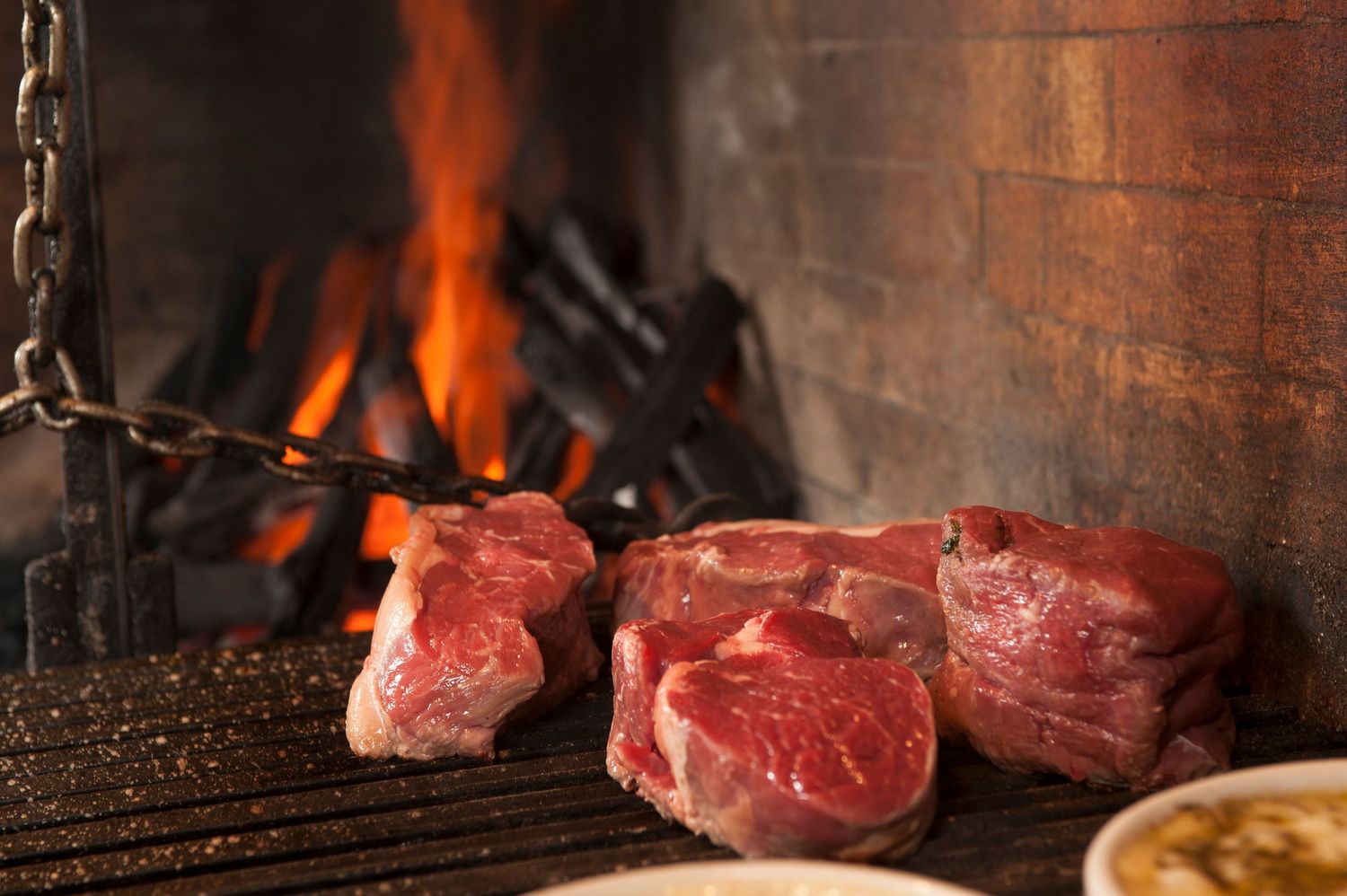 Raw beef cuts grilling over an open fireplace flame on a rustic iron grate in a traditional steakhouse setting.