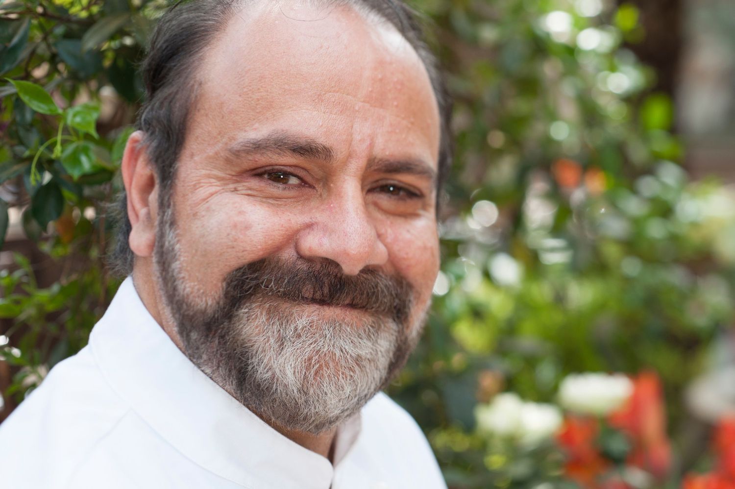 Middle-aged man with beard and salt-and-pepper hair smiling outdoors, wearing white shirt with green foliage background.
