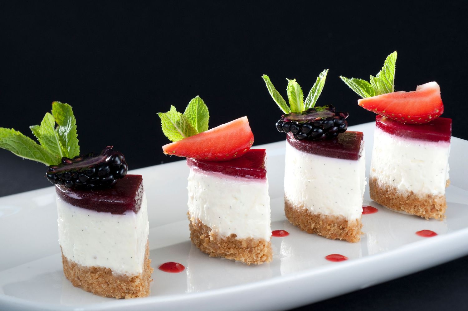 Four elegant mini cheesecakes topped with fresh strawberries, blackberries, and mint leaves on a white plate.