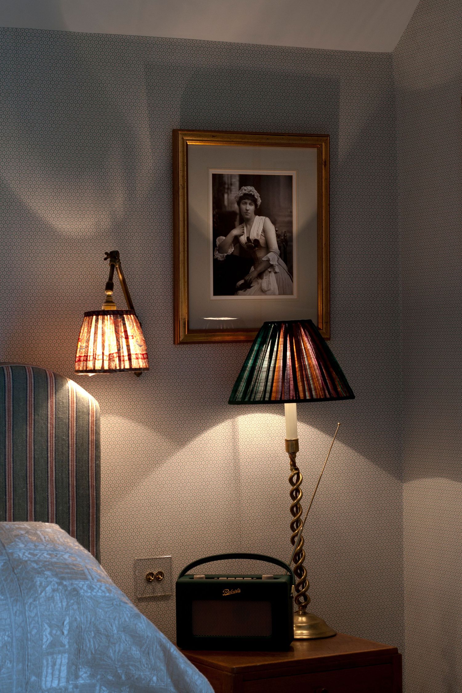 Cozy bedroom corner with wall lamp, table lamp on nightstand, vintage radio, and framed black-and-white portrait photo.
