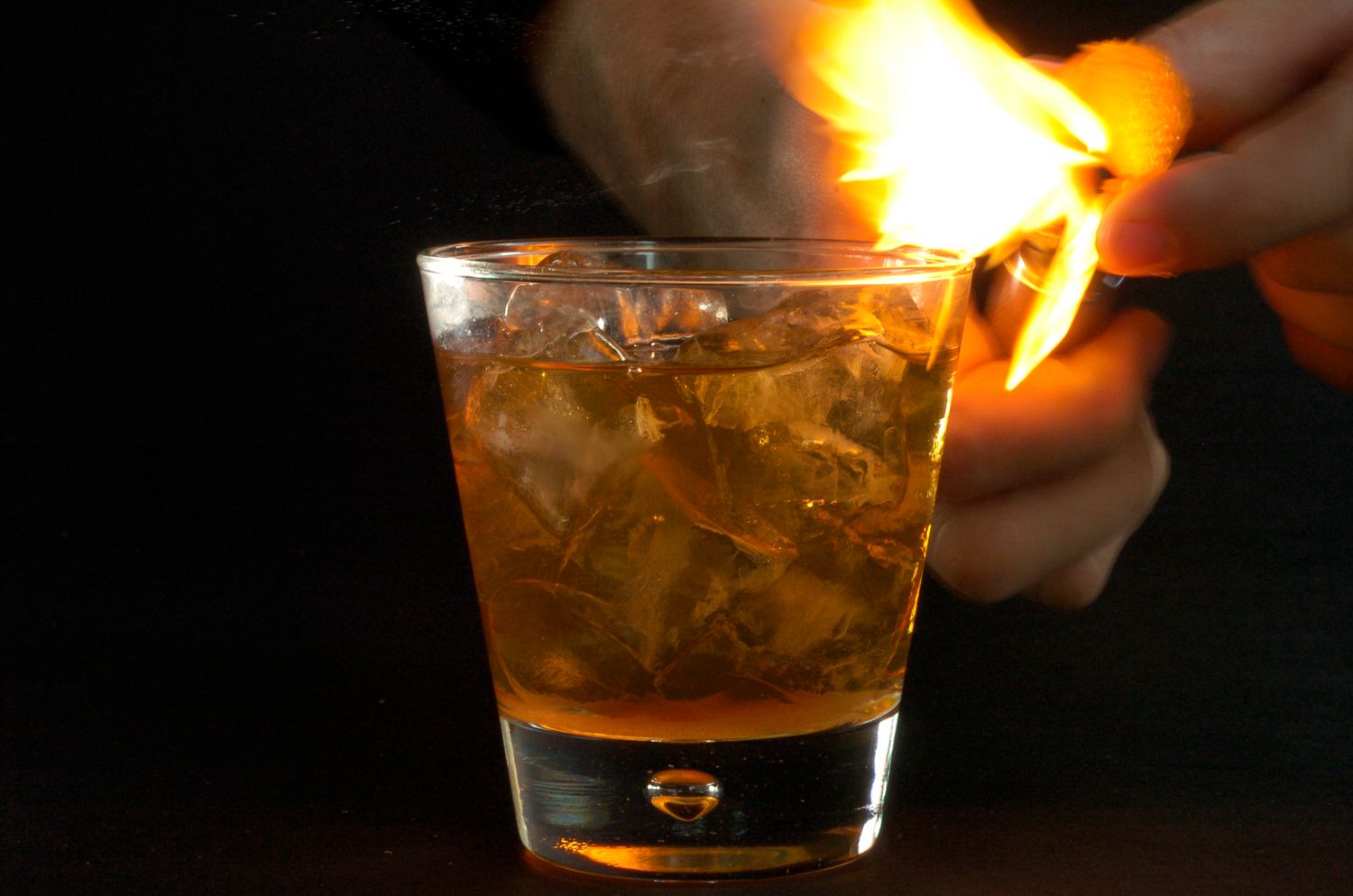 Flaming whiskey cocktail on the rocks in a glass tumbler with hand holding lit match over amber drink.