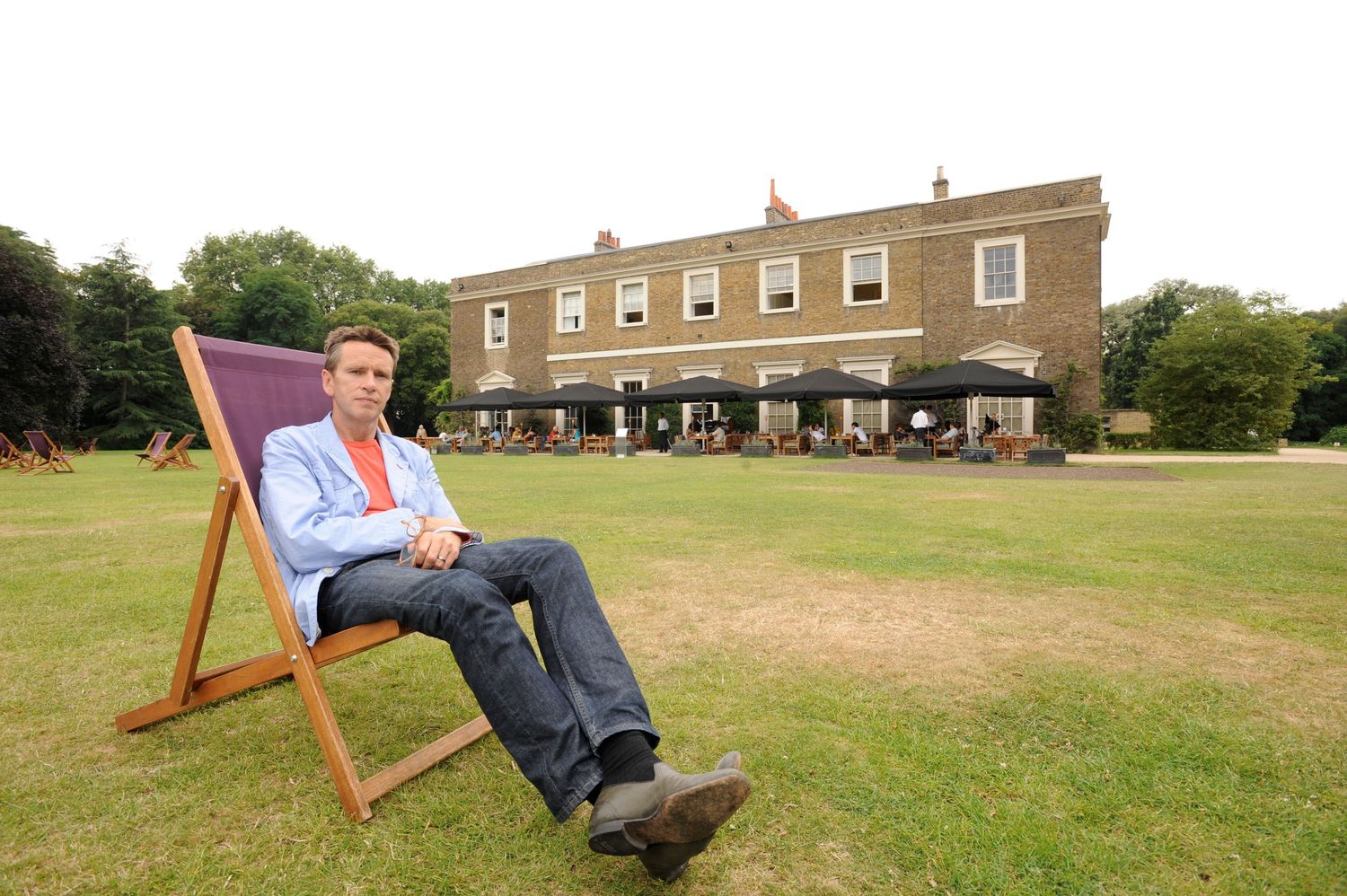 Man relaxing in large wooden deck chair on green lawn outside elegant Georgian country house hotel.