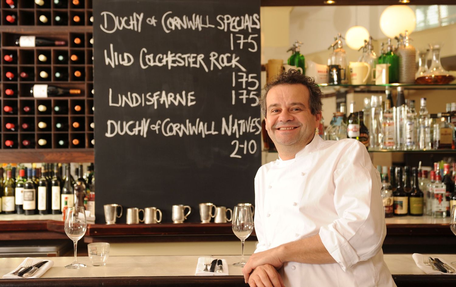 Chef in white uniform smiling in a restaurant with wine rack and specials chalkboard featuring Duchy of Cornwall oysters.
