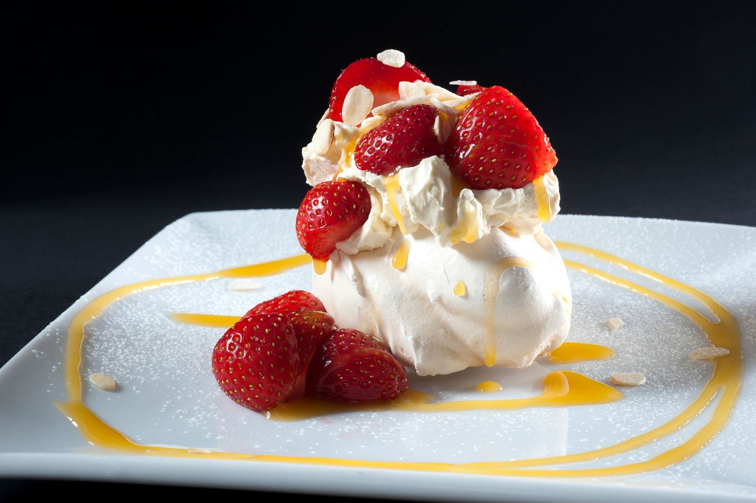 Elegant pavlova dessert topped with fresh strawberries and drizzled with caramel sauce on a white square plate.