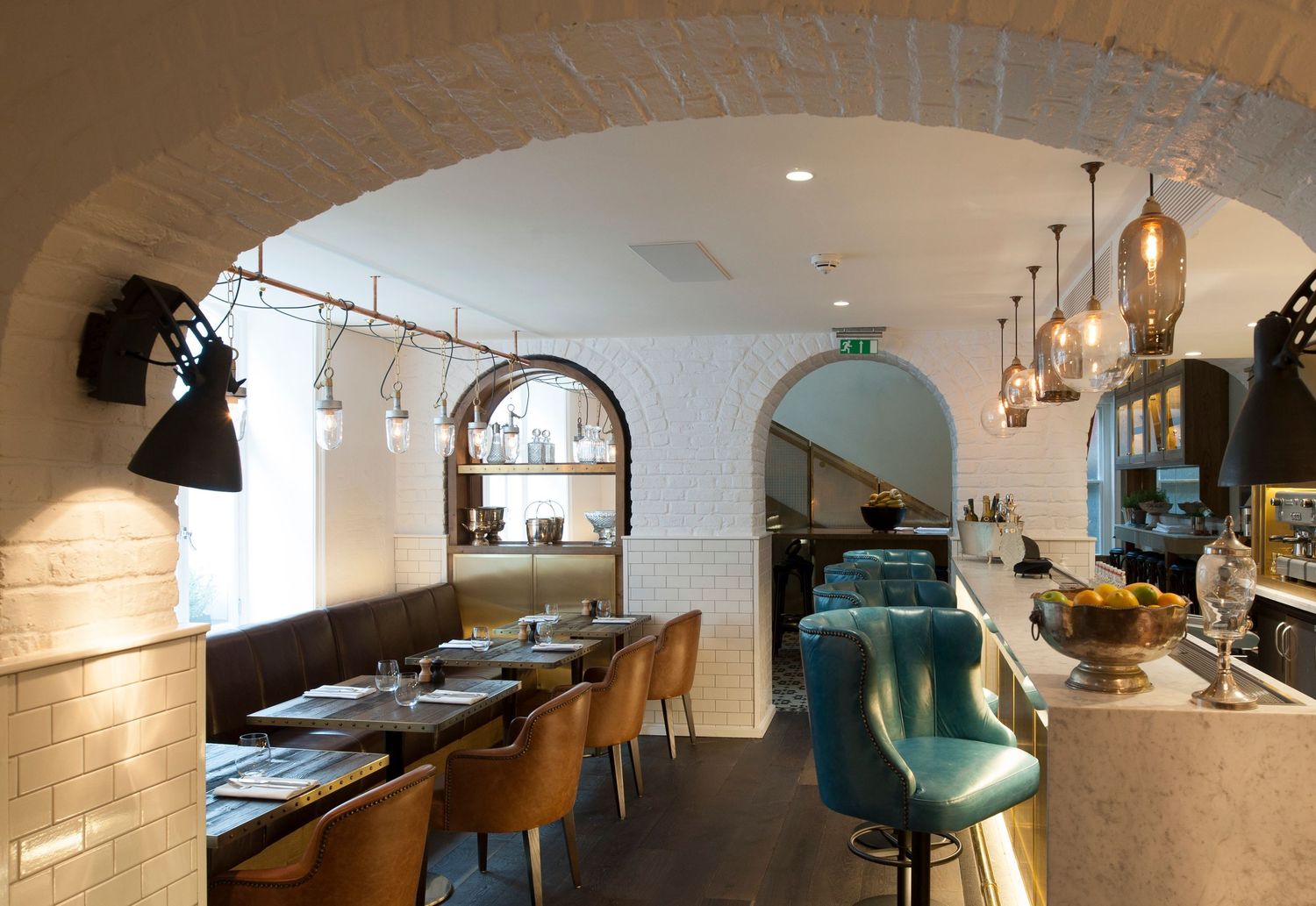 Elegant restaurant interior with arched brick walls, teal bar stools, pendant lights, and wooden dining chairs set with menus.