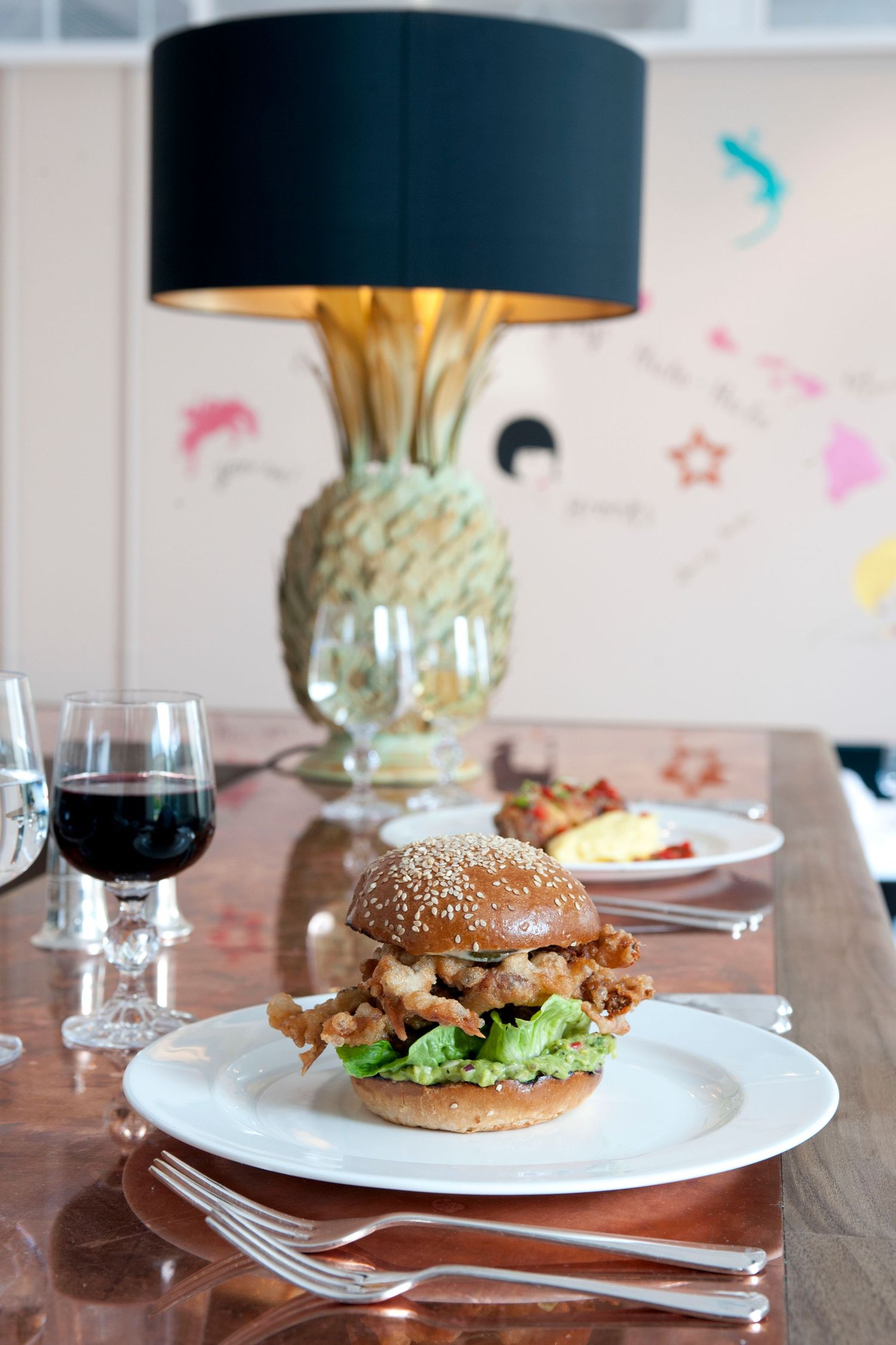 A gourmet burger on a white plate beside a glass of red wine, with a pineapple lamp and artwork in the background.