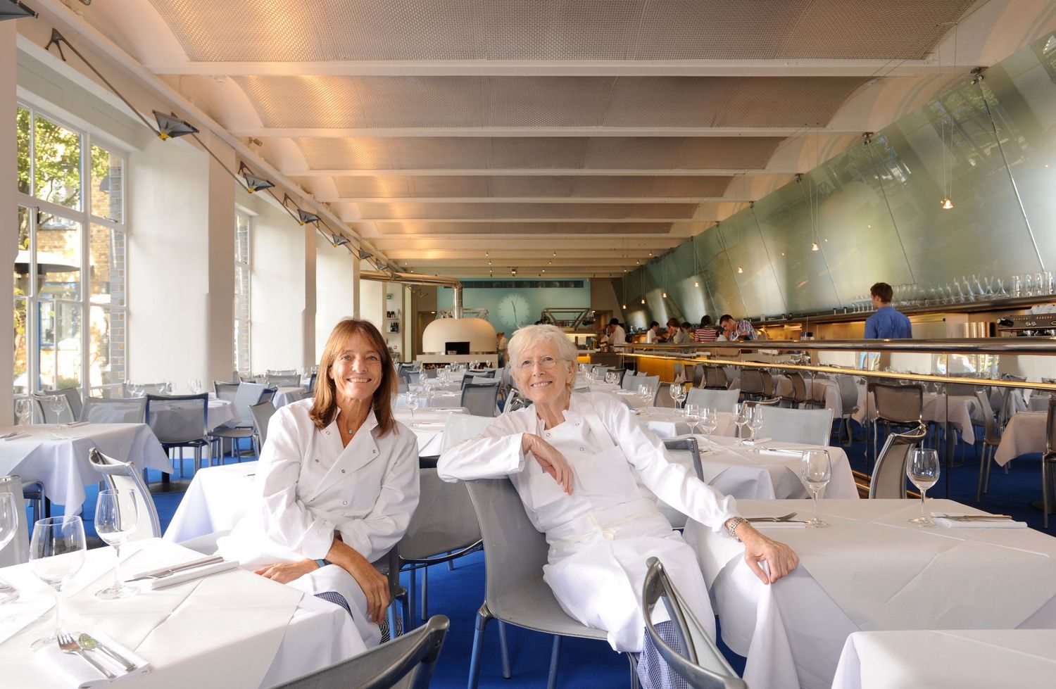 Two people in white attire sit smiling in an elegant restaurant with white tablecloths and blue chairs.