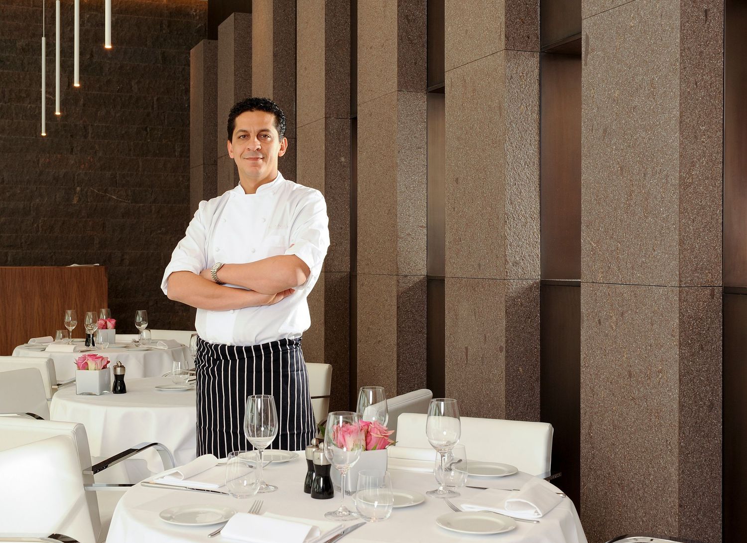 A confident chef in white uniform and striped apron stands arms crossed in an elegant restaurant dining room.