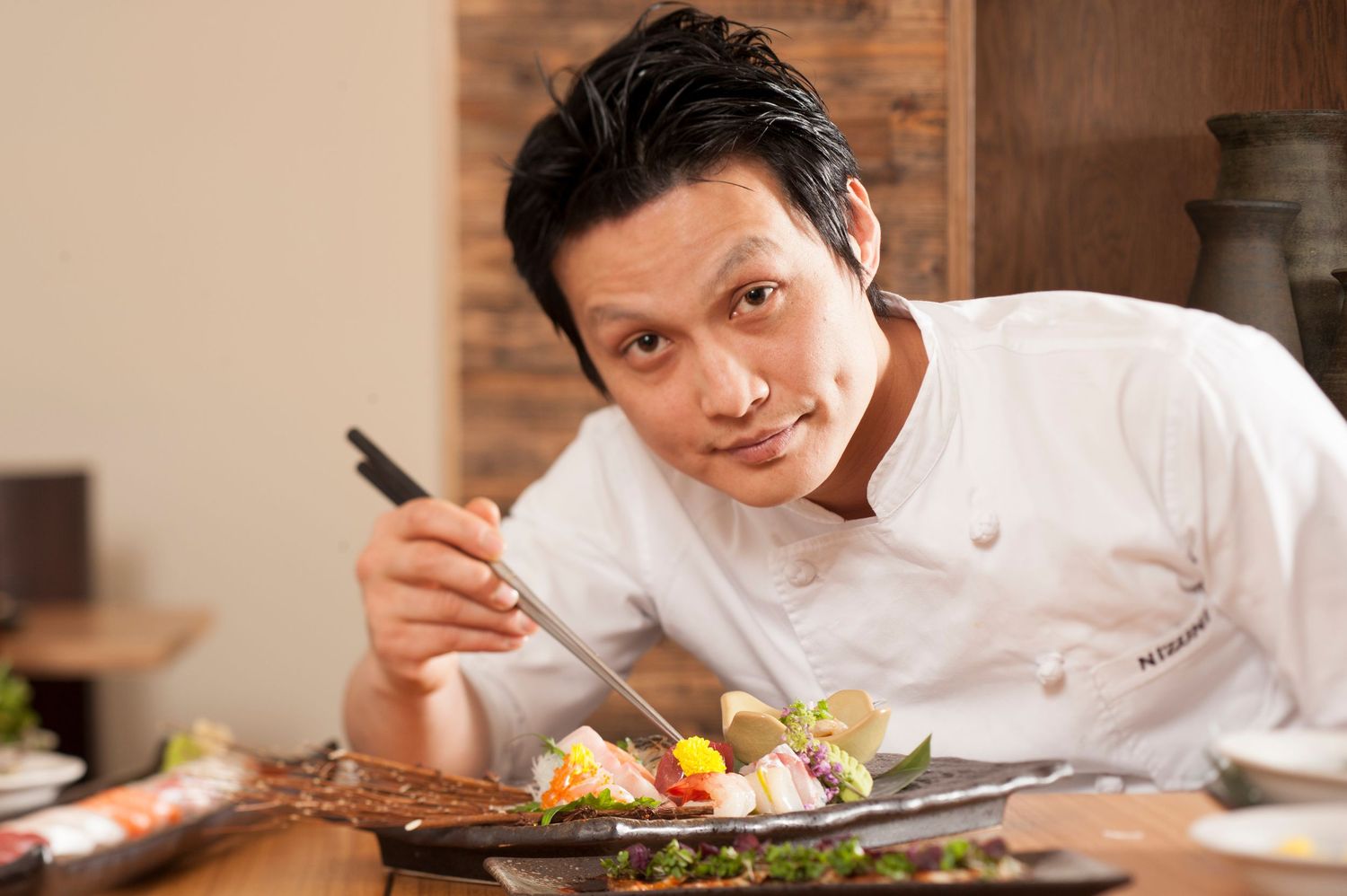 A smiling Asian chef in white uniform carefully plates a colorful gourmet dish with chopsticks in a restaurant.