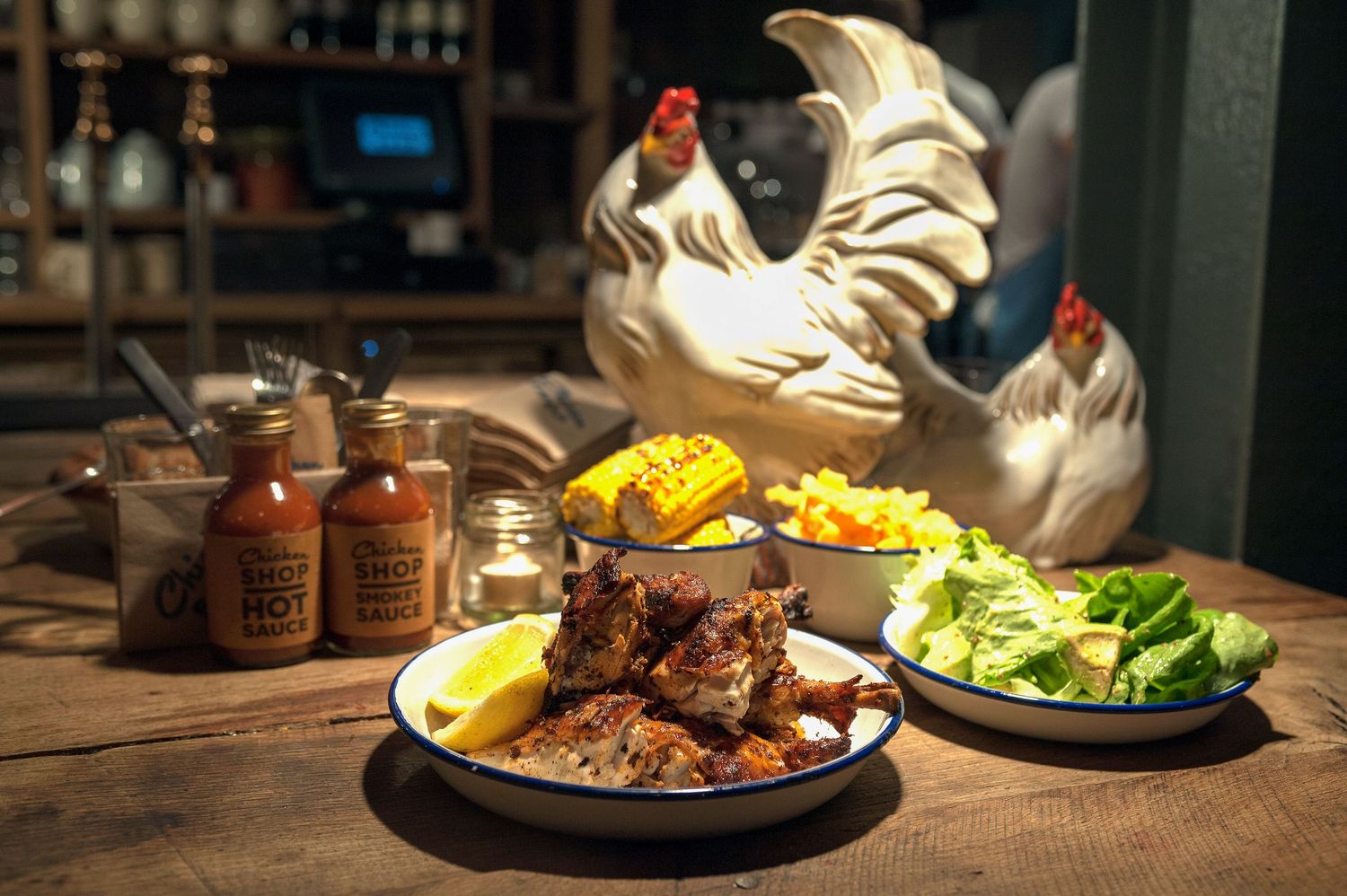 BBQ ribs with corn, lemon, and fresh salad served on a rustic table beside decorative rooster figurines.