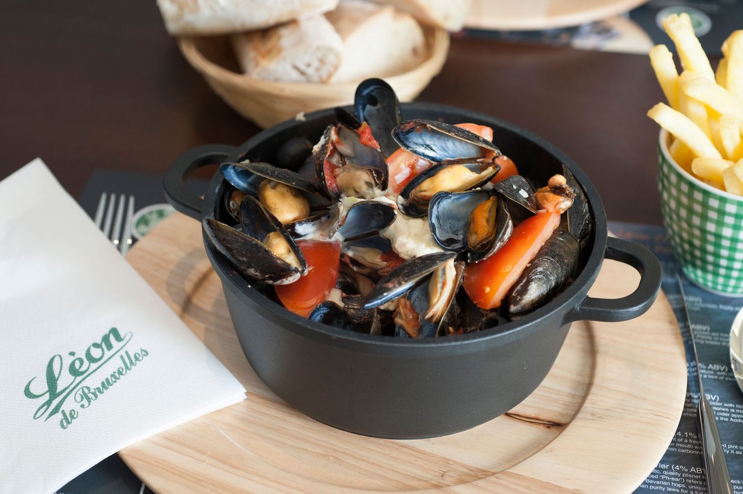 Leon de Bruxelles mussels served in a black cast iron pot with carrots, accompanied by golden French fries on a wooden board.
