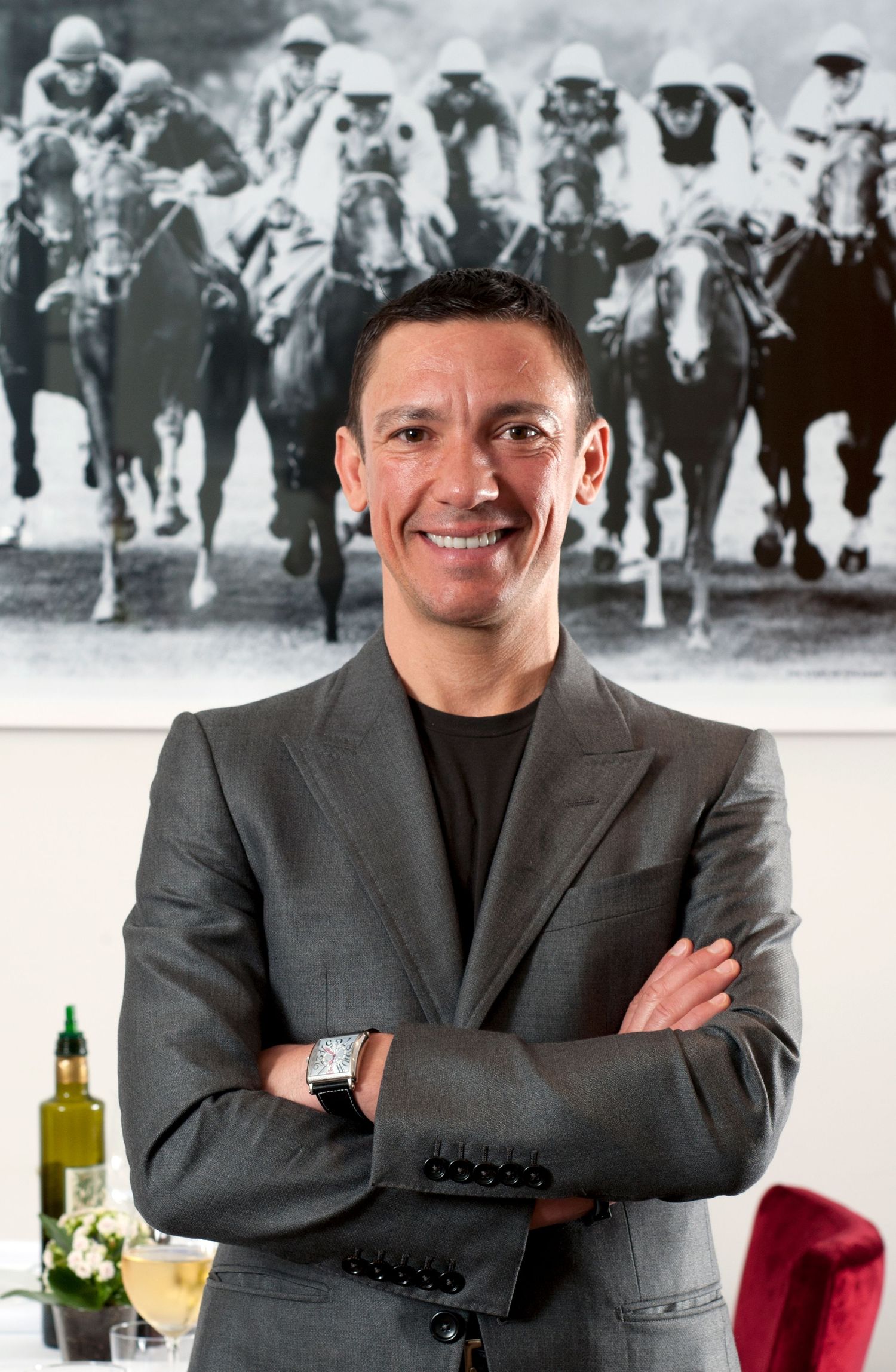 Smiling man in gray blazer with arms crossed stands before a black and white horse racing photograph.