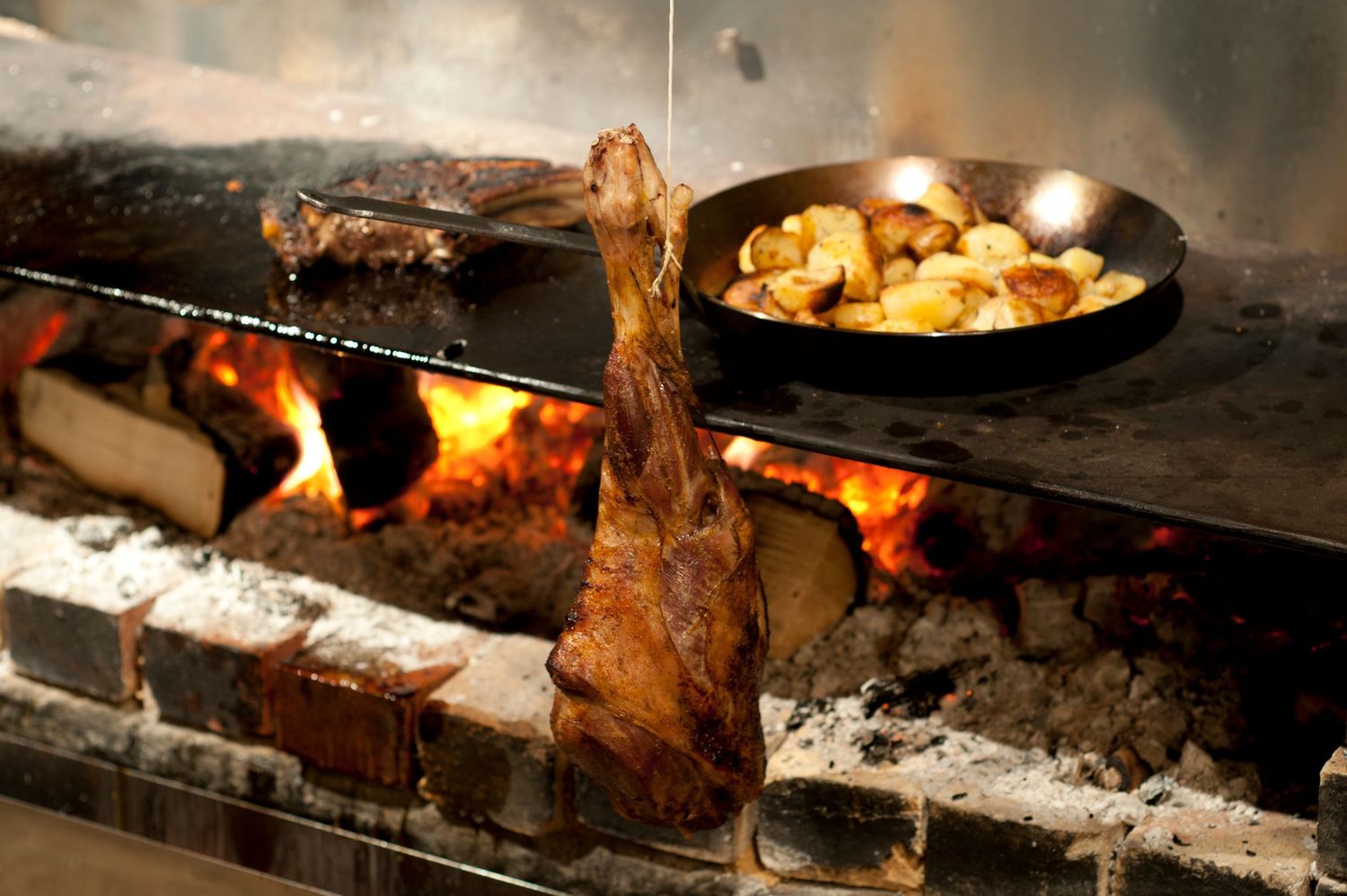 Lamb leg roasting over open wood fire with golden potatoes cooking in cast iron skillet at rustic steakhouse.
