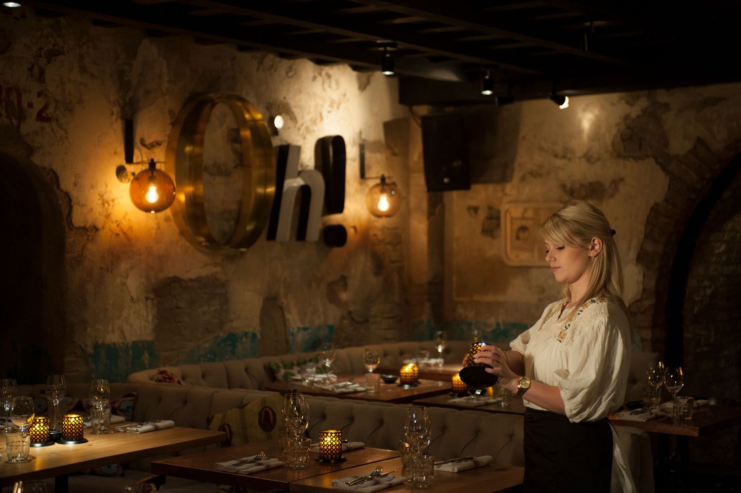 A woman stands in a dimly lit rustic restaurant with exposed stone walls, warm Edison bulbs, and an elegantly set dining table.