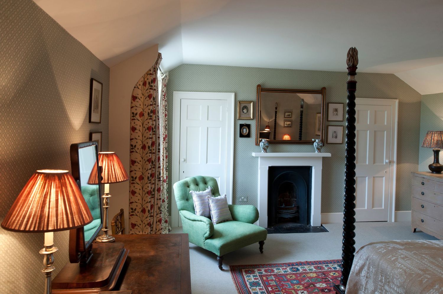 Classic bedroom with four-poster bed, fireplace, green armchair, and warm lamp lighting on hardwood floors.