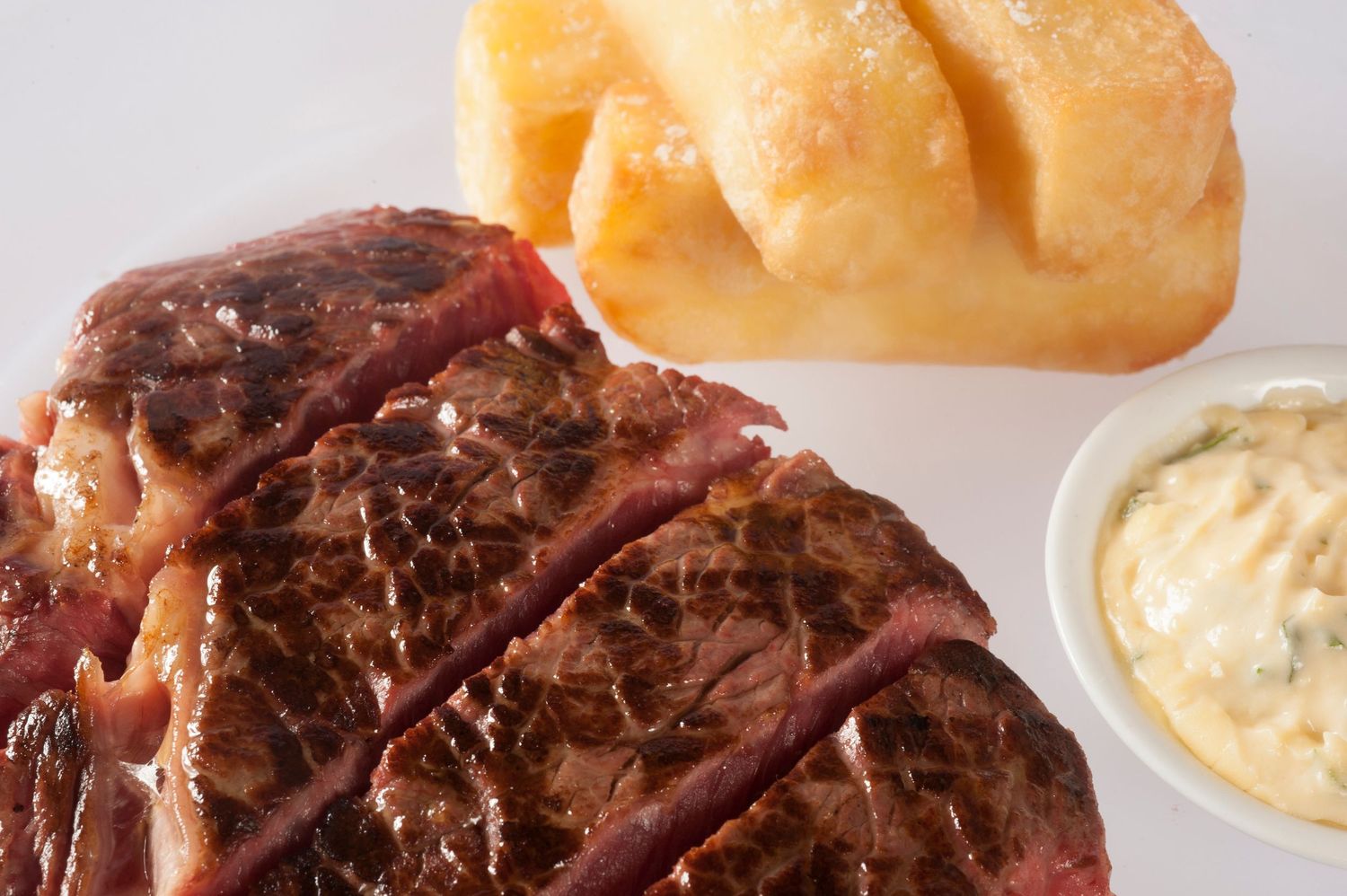 Juicy grilled ribeye steak served with golden fried potatoes and a side of creamy tartar sauce on a white plate.