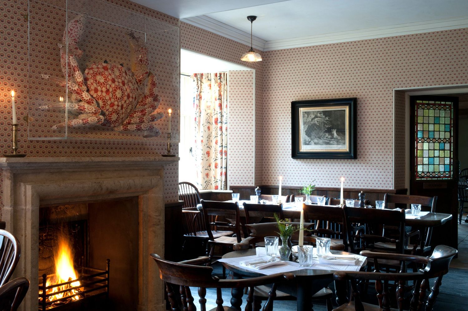 Cozy restaurant dining room with lit fireplace, dark wood chairs, set tables, patterned wallpaper, and stained glass window.