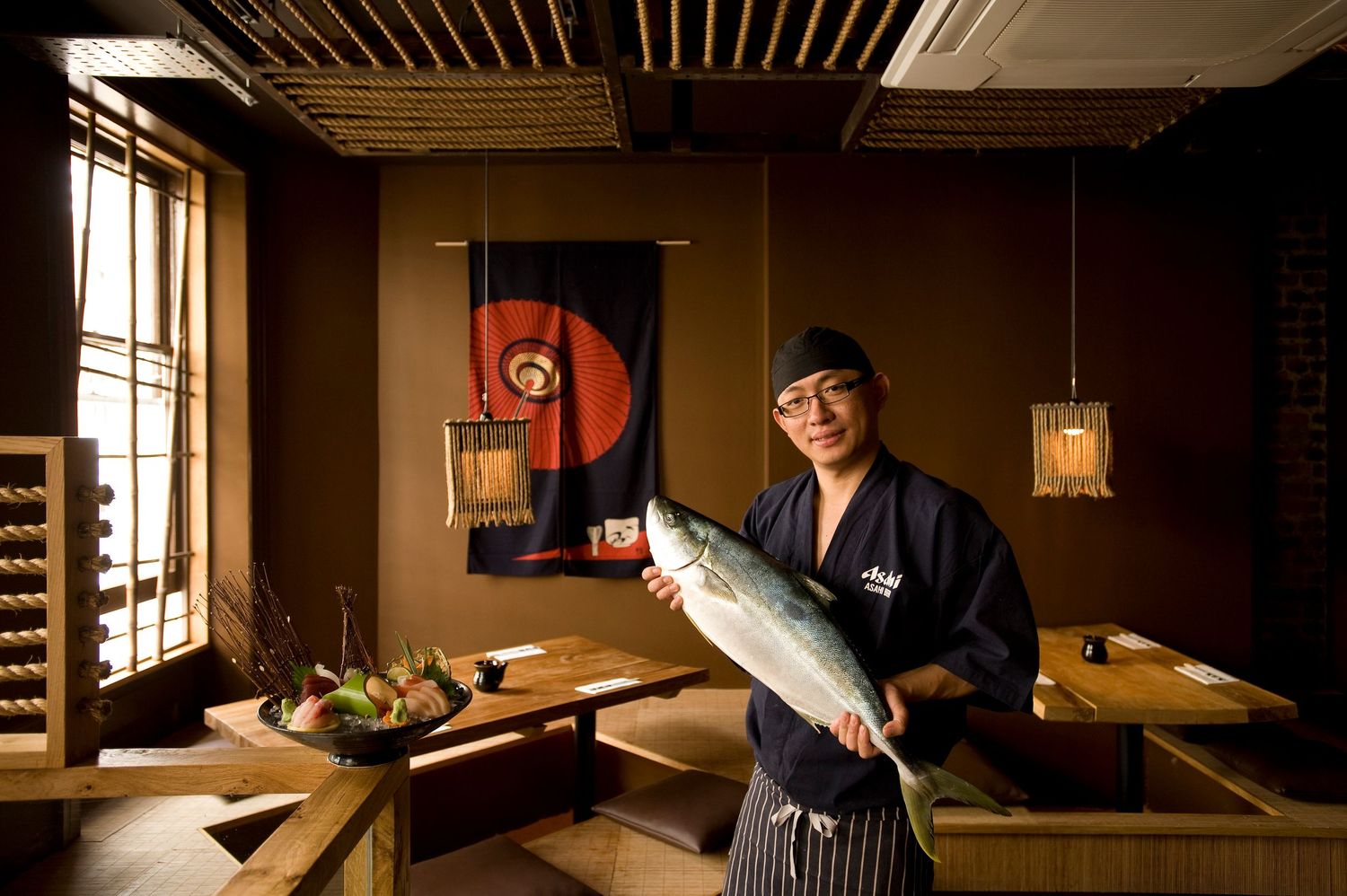 Japanese chef proudly holding a fresh whole fish in a traditional Japanese restaurant with wooden decor.