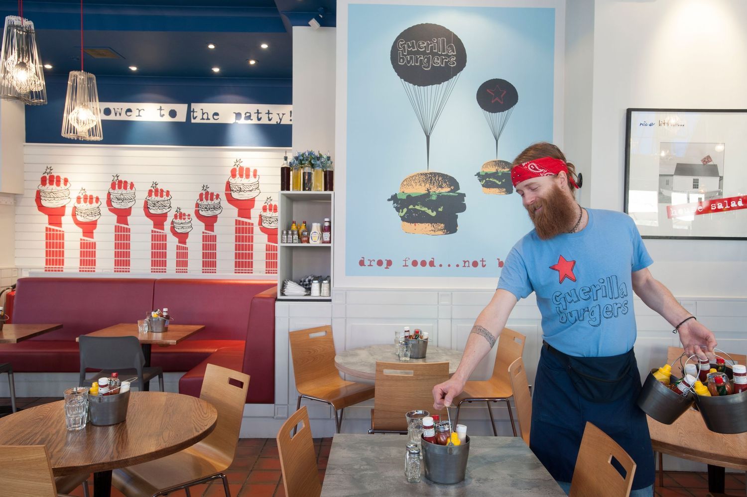 A bearded man in a blue t-shirt sets tables in a vibrant burger restaurant with colorful wall murals.