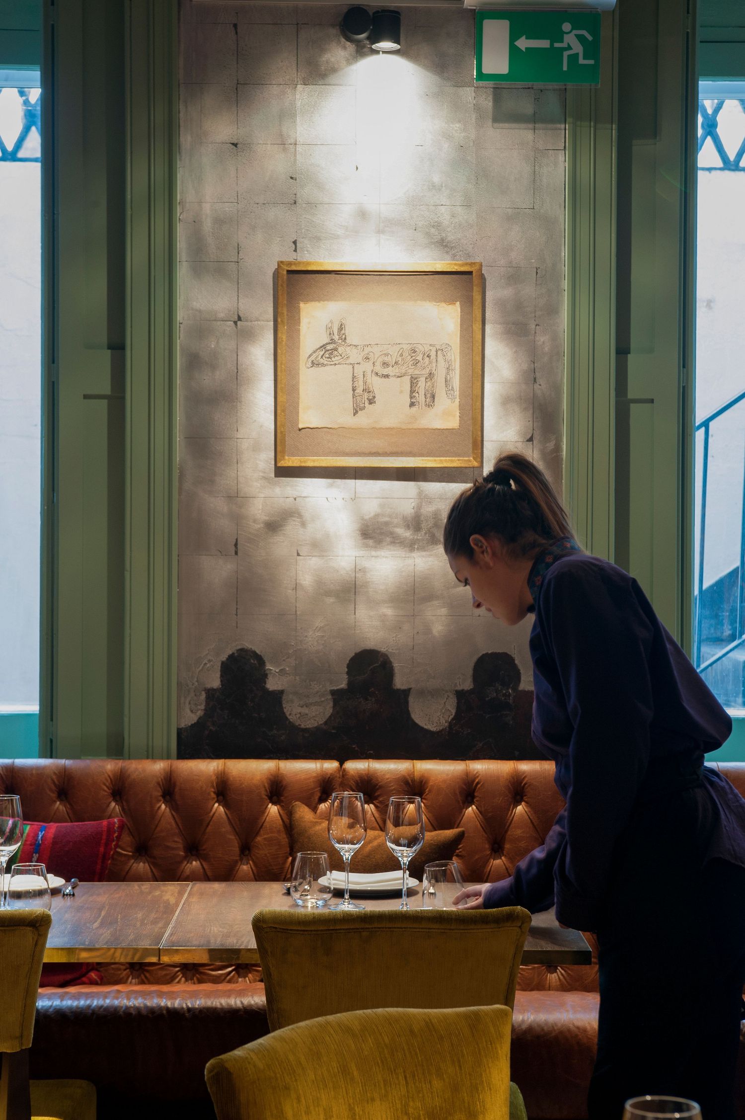 A waitress sets wine glasses on a leather banquette table in an elegant restaurant with framed artwork on the wall.