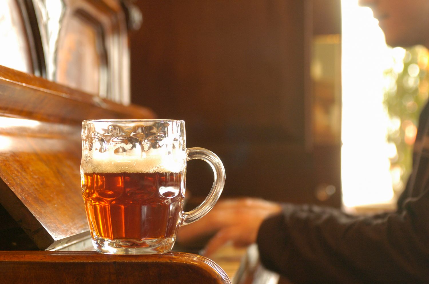 A frothy glass mug of amber beer resting on a wooden bar counter in a warm, sunlit pub.