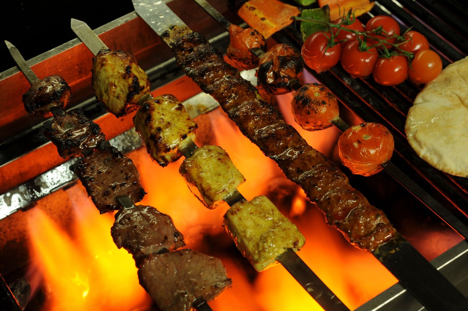 Skewers of grilled meat and tomatoes cooking over glowing orange charcoal on a BBQ grill.