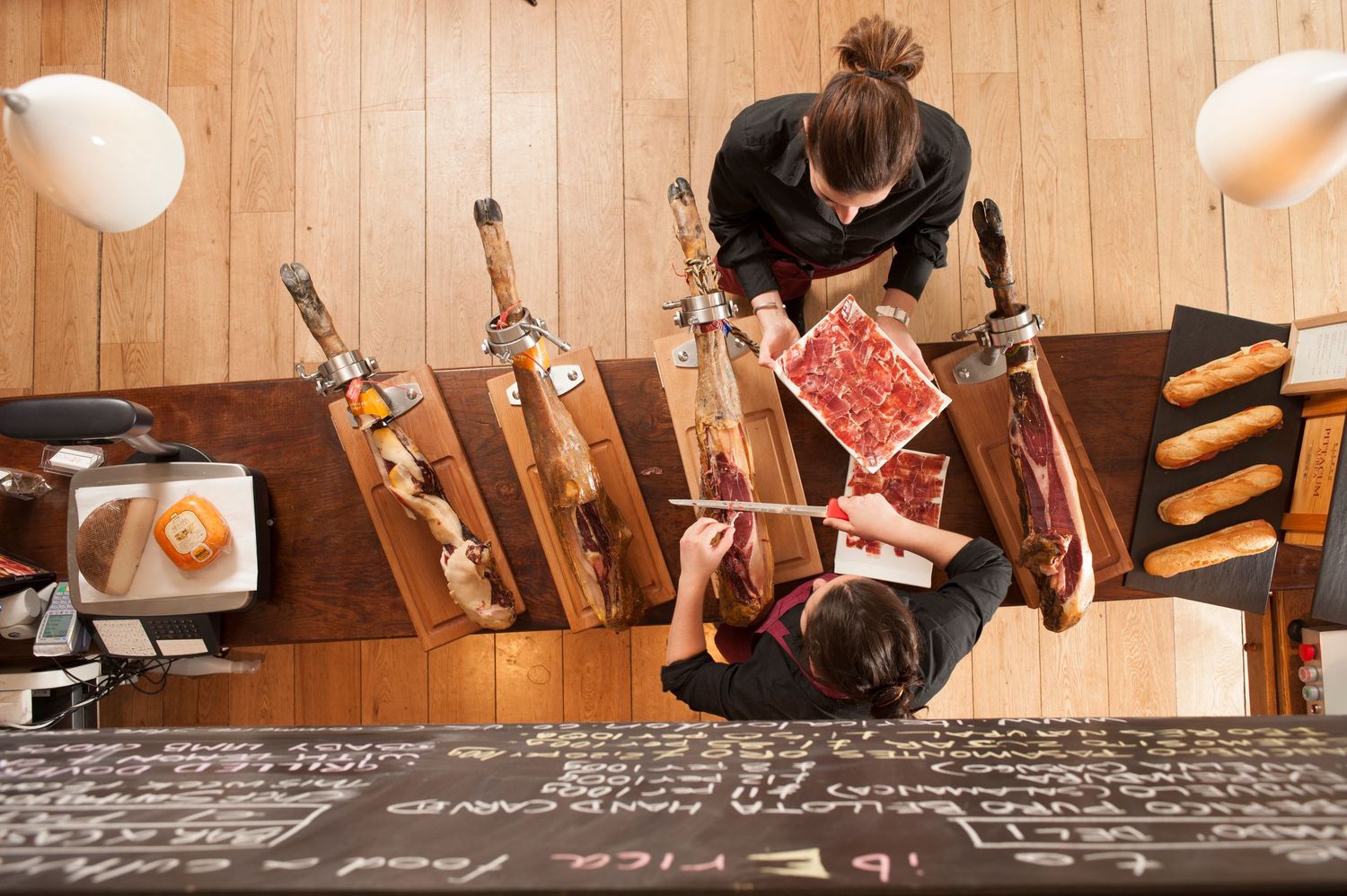 Aerial view of a Spanish tapas bar with jamón legs on stands and staff serving charcuterie on a chalkboard counter.