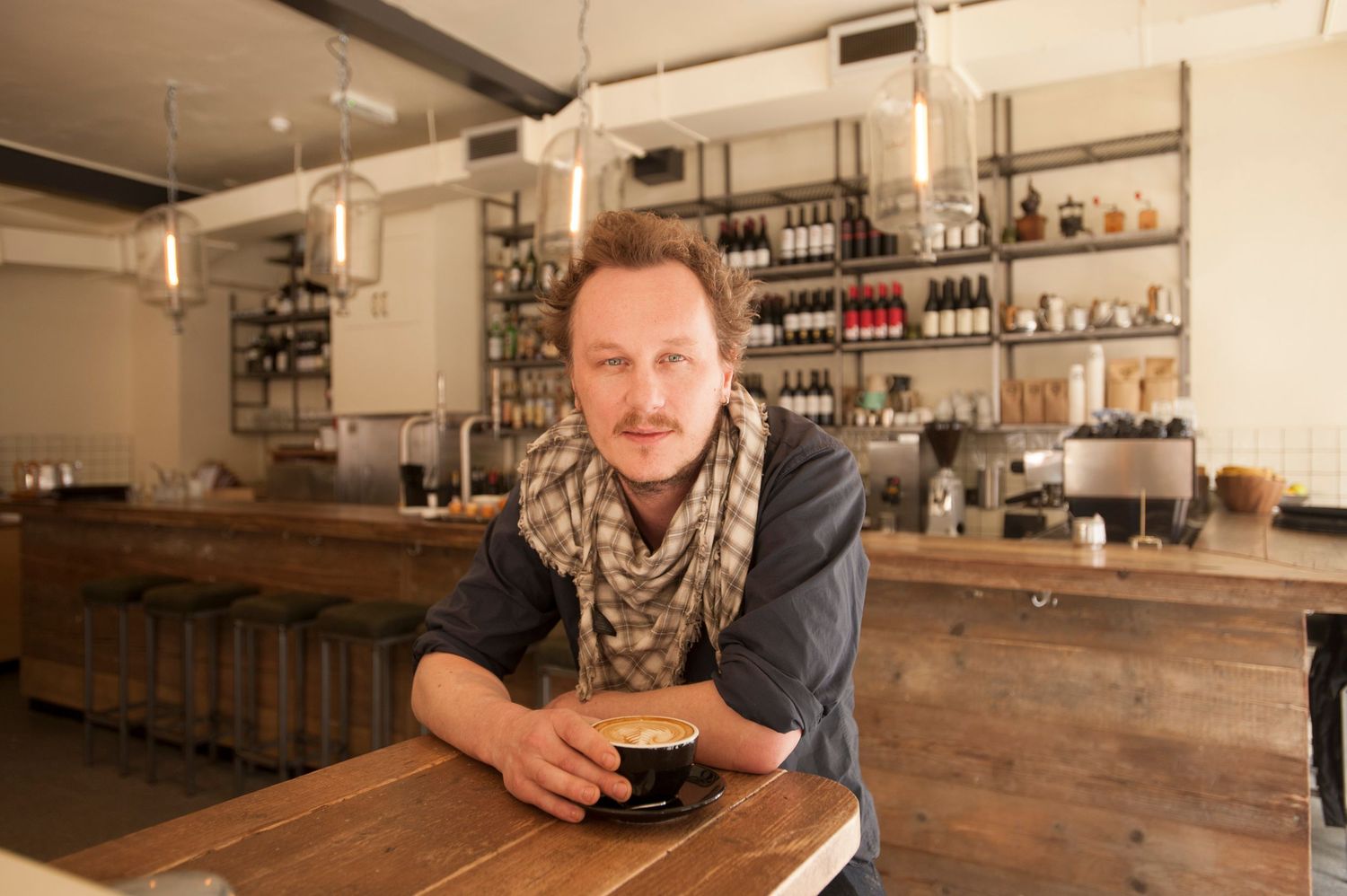 Man with scarf holding a coffee cup leaning on a wooden bar counter in a modern cafe with bottle-lined shelves.