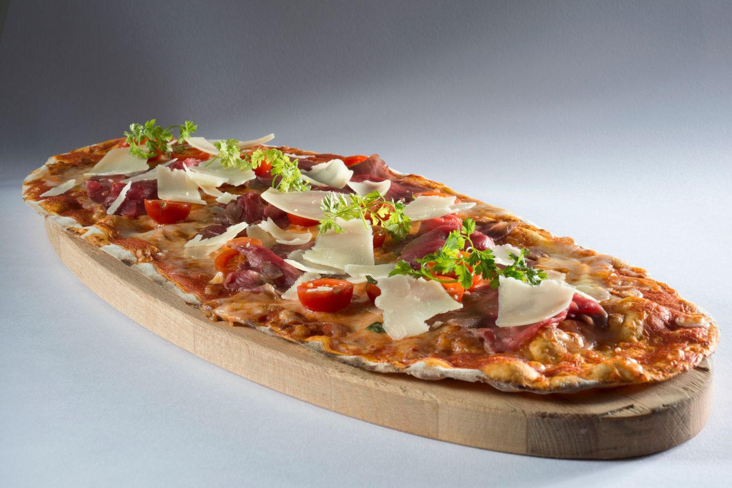 Oval flatbread pizza topped with prosciutto, cherry tomatoes, shaved parmesan, and fresh greens on a wooden board.
