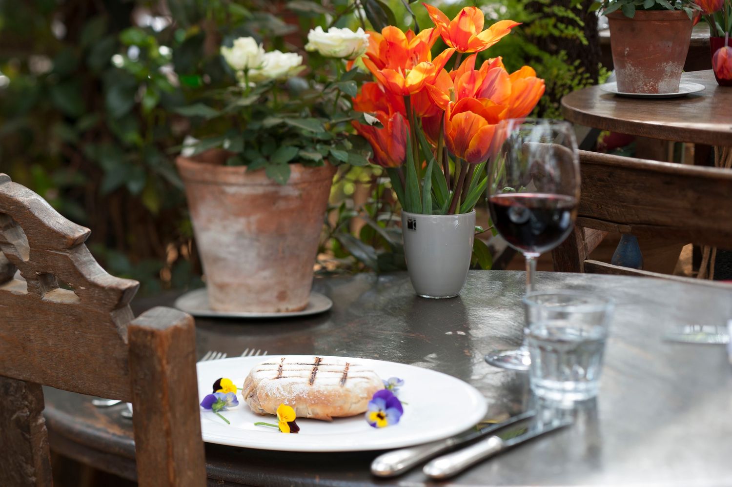 Outdoor dining table with a gourmet dish garnished with edible flowers, red wine, water glass, and vibrant orange tulips in a vase.