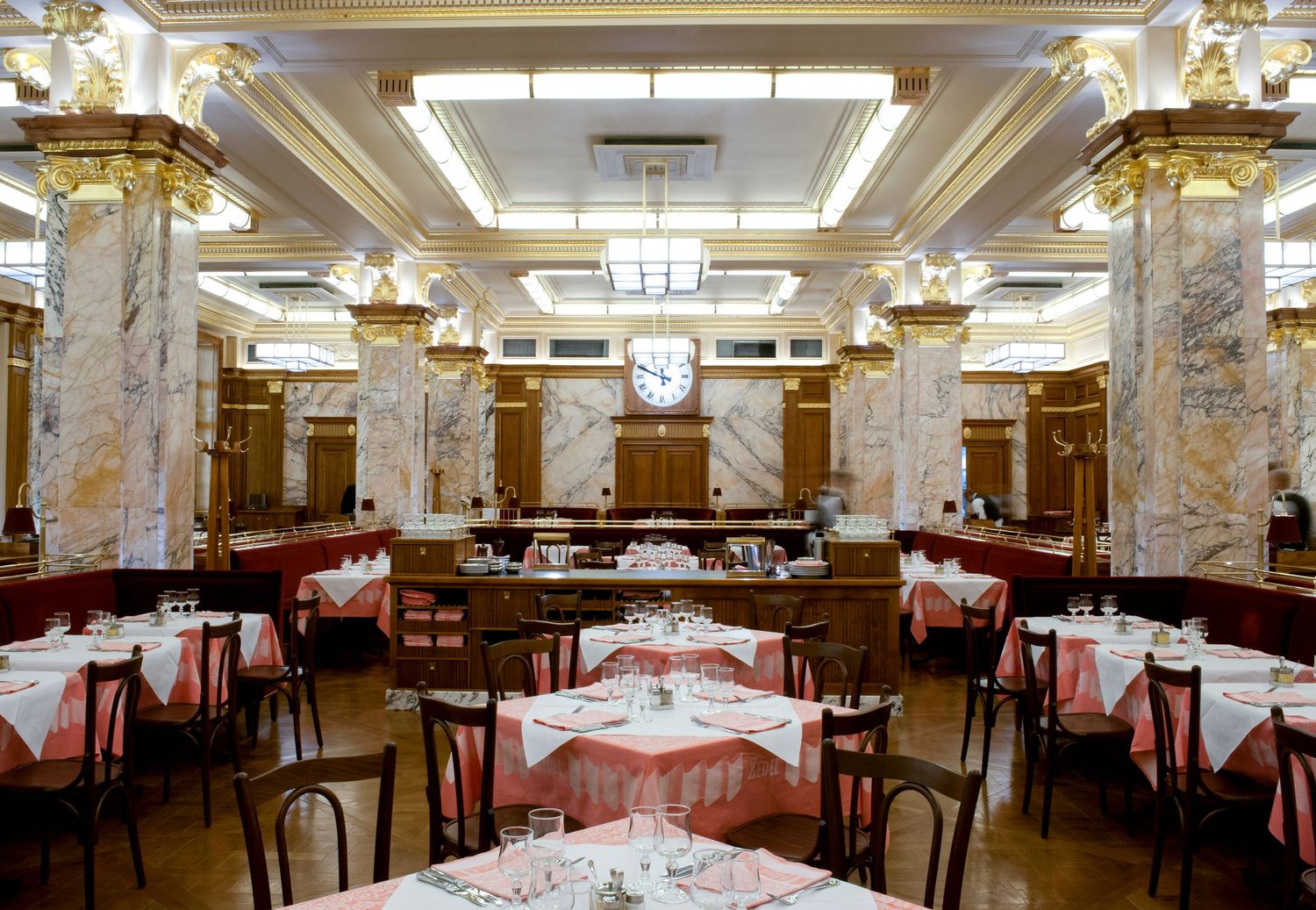 Elegant restaurant interior with marble columns, gold accents, red tablecloths, and a classic wall clock.