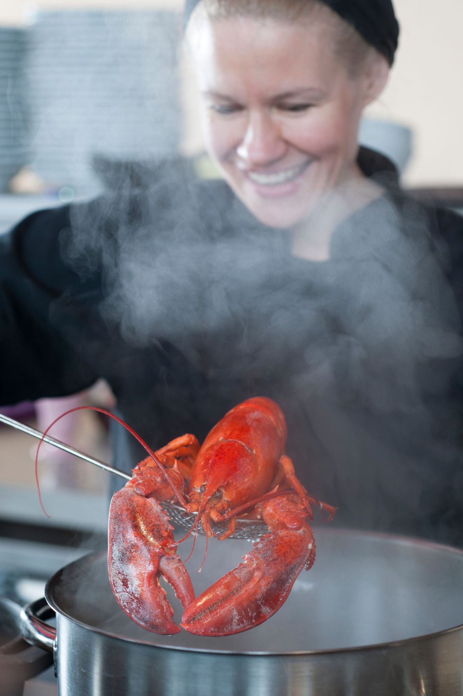 Smiling female chef lifting a bright red steamed lobster from a steaming pot using tongs in a professional kitchen.