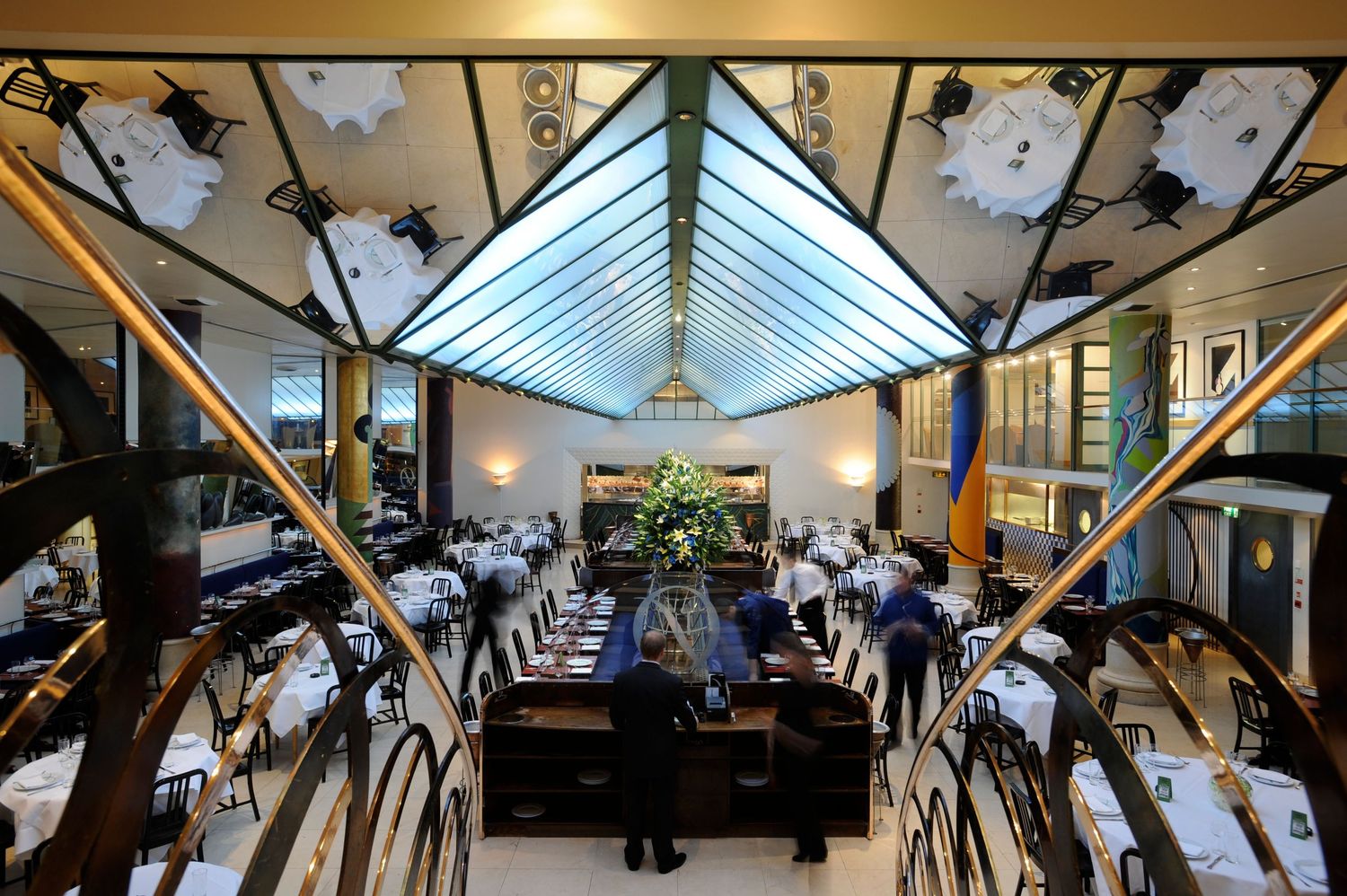 Elegant restaurant interior with skylight ceiling, white tablecloths, and staff serving guests in a grand dining hall.