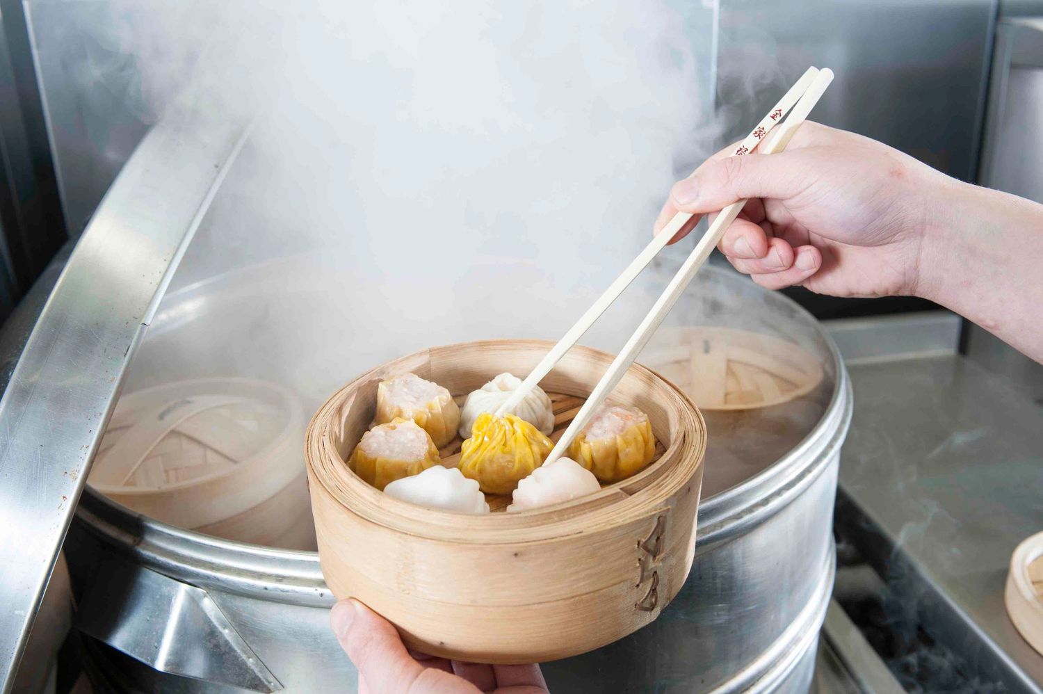 Hands using chopsticks to pick up dim sum dumplings from a bamboo steamer basket surrounded by steam.