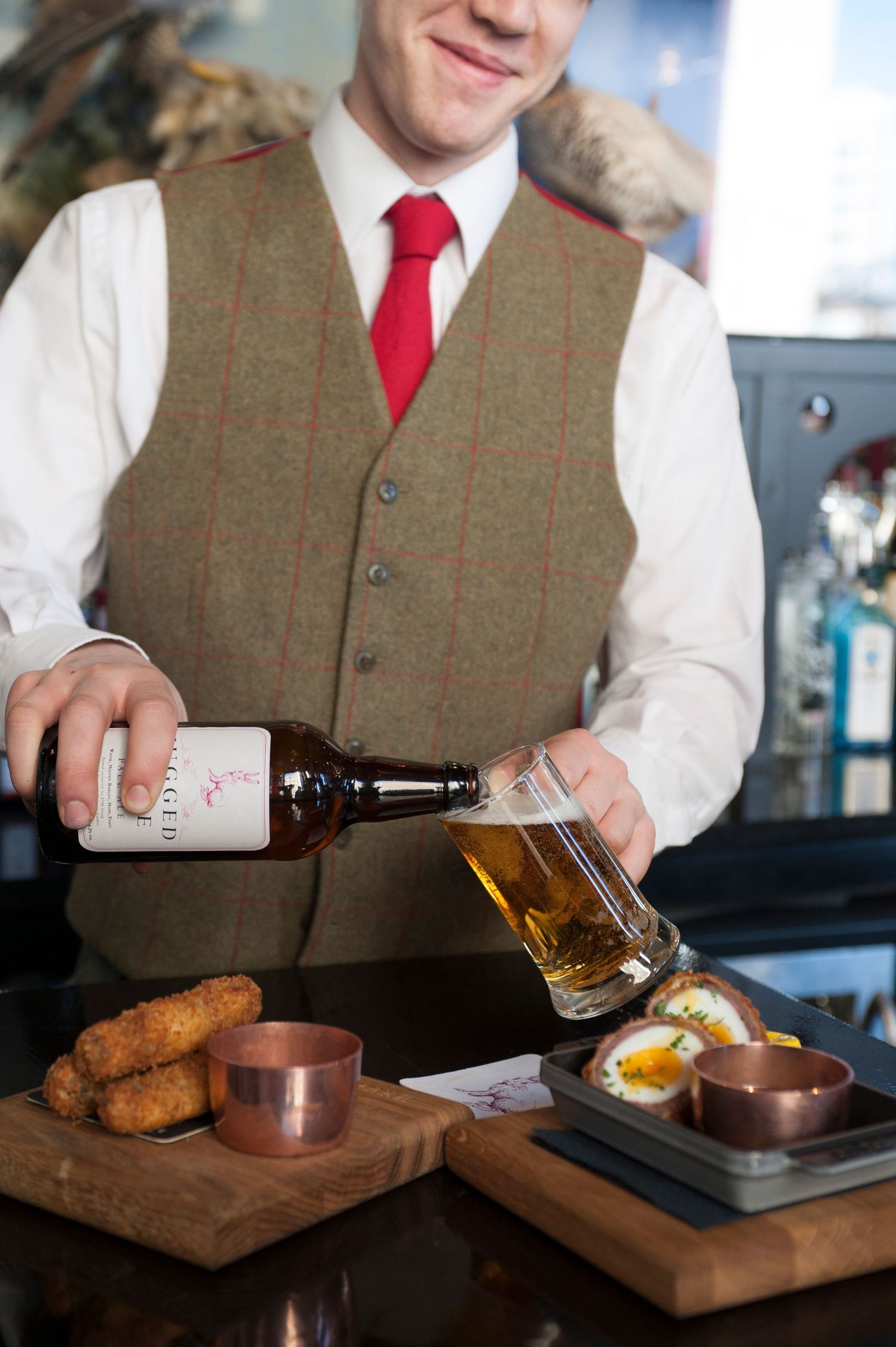Bartender in tweed vest and red tie pouring craft beer into glass pint beside scotch eggs and croquettes.