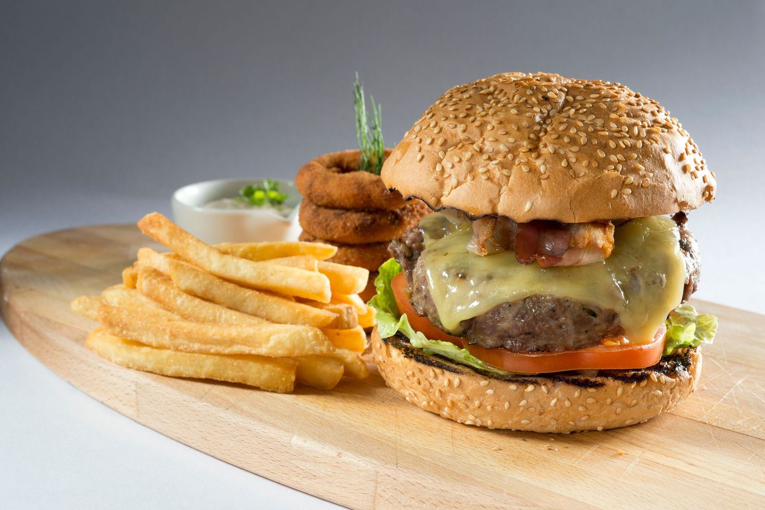 Juicy cheeseburger with bacon, onion rings, and fresh veggies on sesame bun served with crispy fries on wooden board.