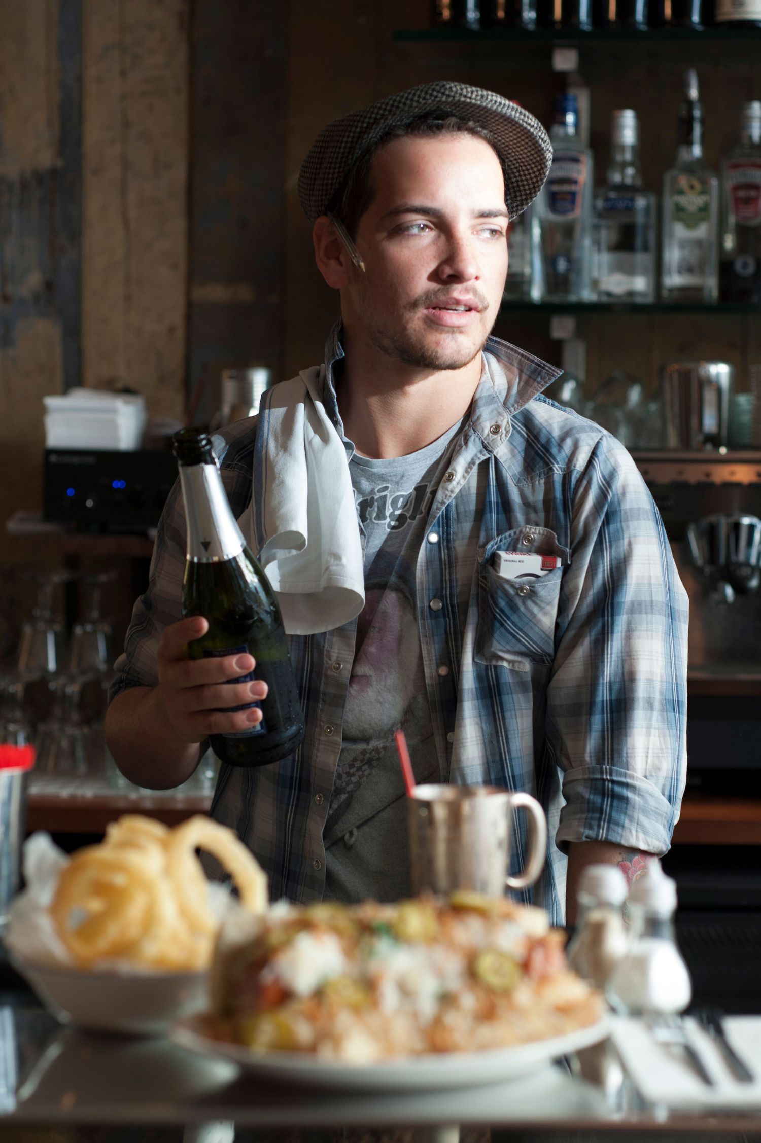 A bartender in a plaid shirt and hat holds a wine bottle and glass in a bar setting with food in the foreground.