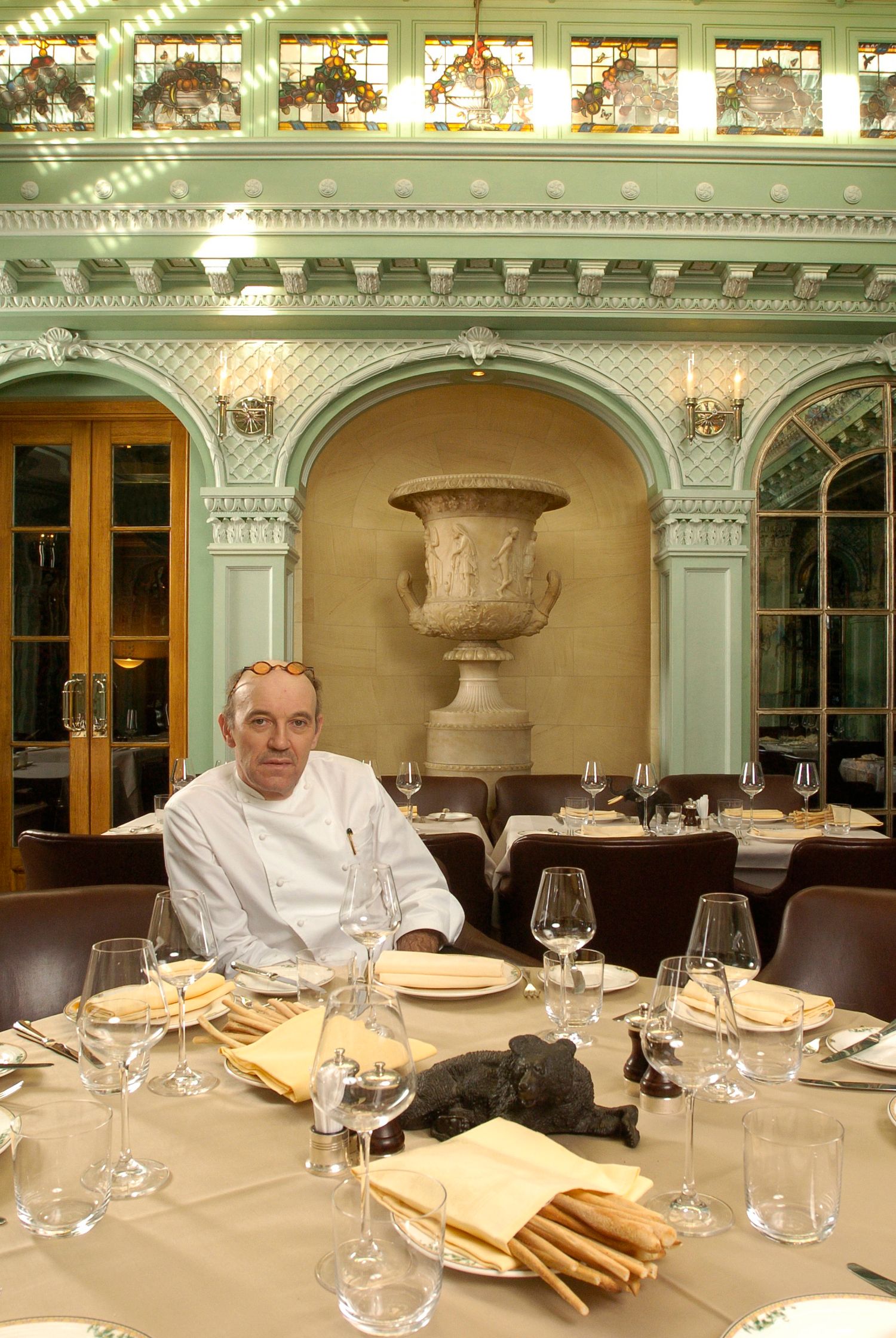 Chef in white uniform seated at elegantly set dining table in ornate restaurant with green arched decor.