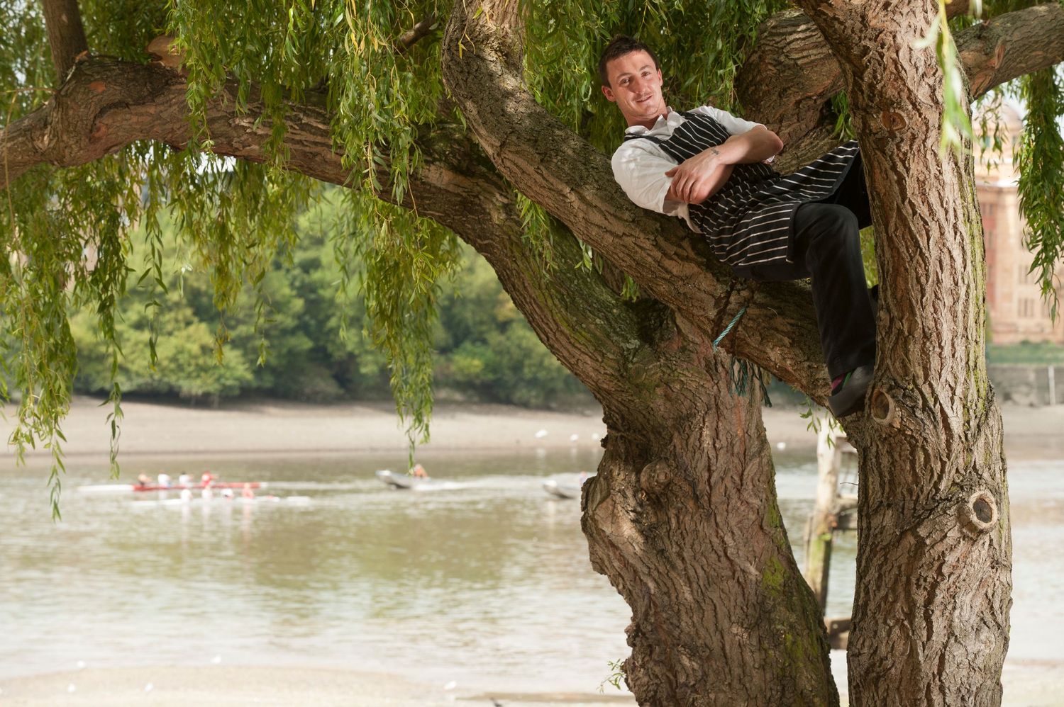 Man relaxing in a large willow tree by a river, with rowing boats visible on the water in the background.