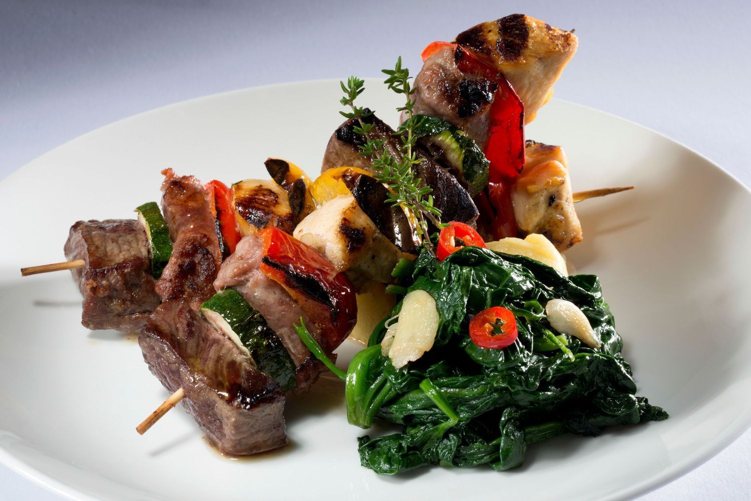 Grilled meat and vegetable skewers served with sautéed spinach and garlic on a white plate.