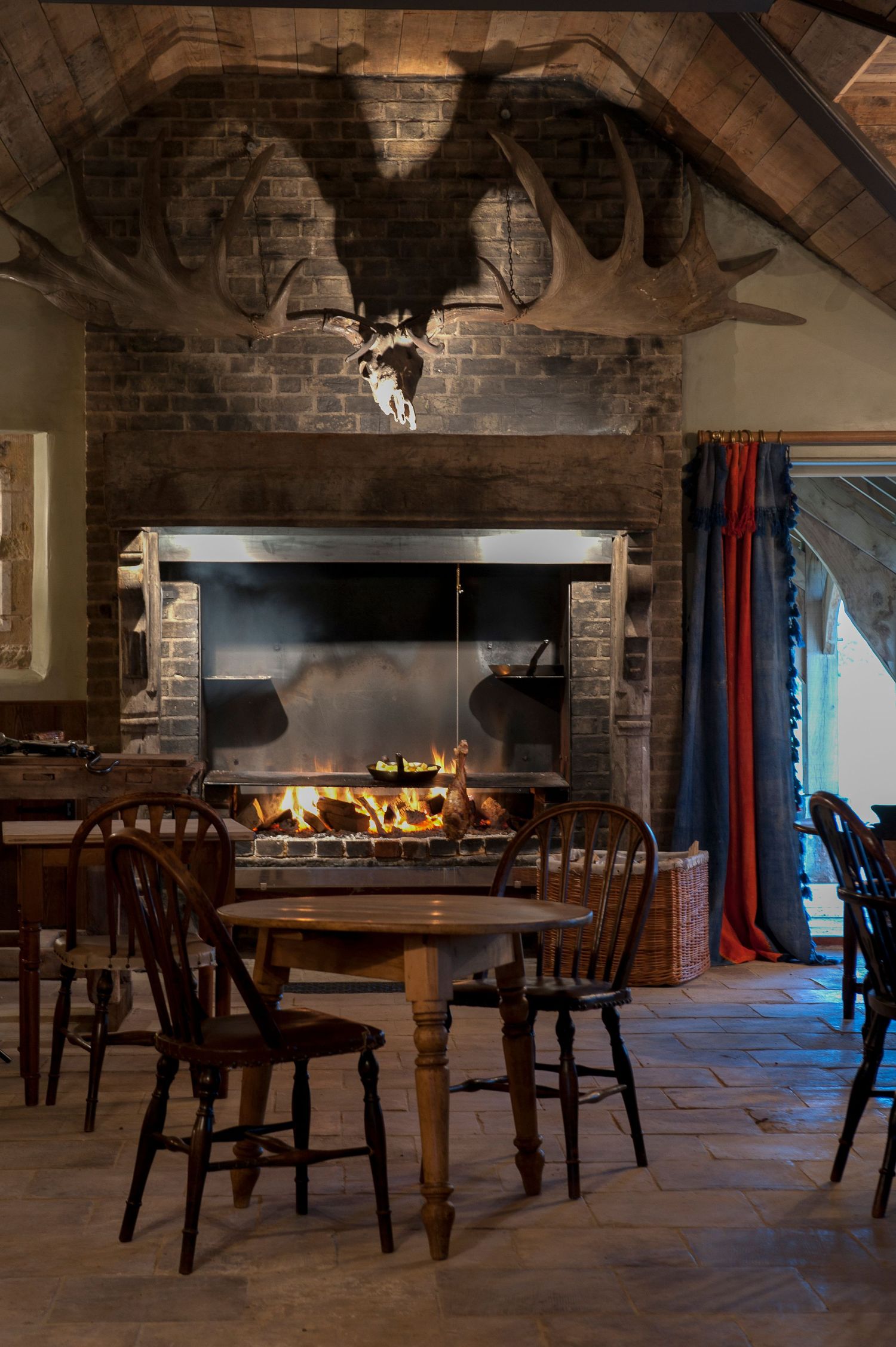 Rustic dining room with wooden chairs, round table, open fireplace, brick walls, and mounted moose antlers on vaulted ceiling.
